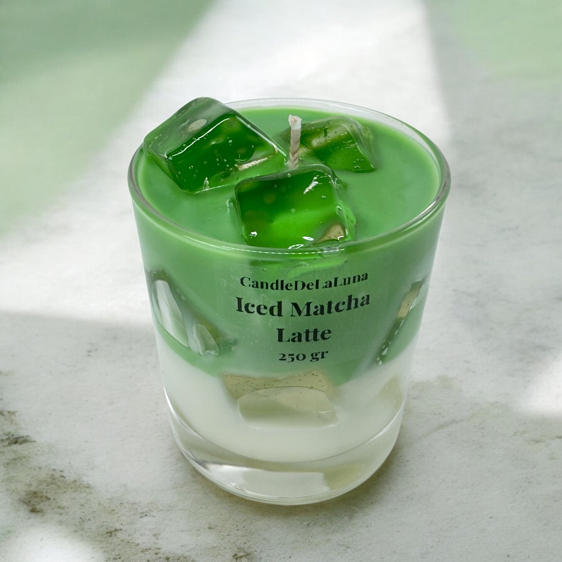 Eine handgemachte Kerze im Design von Iced Matcha Latte in einem niedrigen Glas mit grün-weißen Wachsschichten, künstlichen grünen Eiswürfeln auf der Oberseite und der Aufschrift „CandleDeLuna Iced Matcha Latte 250 gr“, platziert auf einer hellen Marmoroberfläche unter natürlichem Lichteinfall.