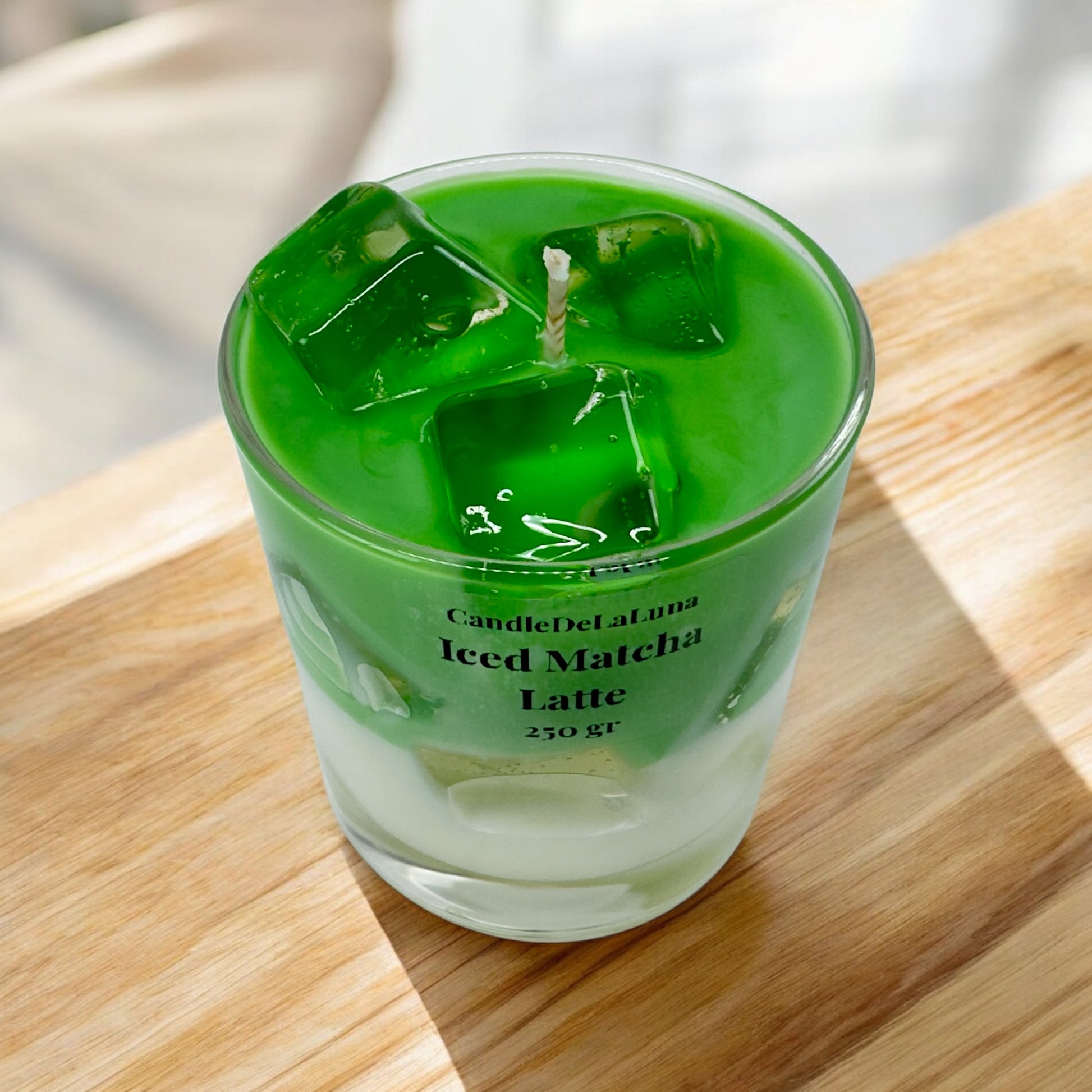Eine handgemachte Kerze im Design von Iced Matcha Latte in einem niedrigen Glas mit grün-weißen Wachsschichten, künstlichen grünen Eiswürfeln aus Wachs auf der Oberseite und der Aufschrift „CandleDeLuna Iced Matcha Latte 250 gr“, platziert auf einer hellen Holzoberfläche im Sonnenlicht.