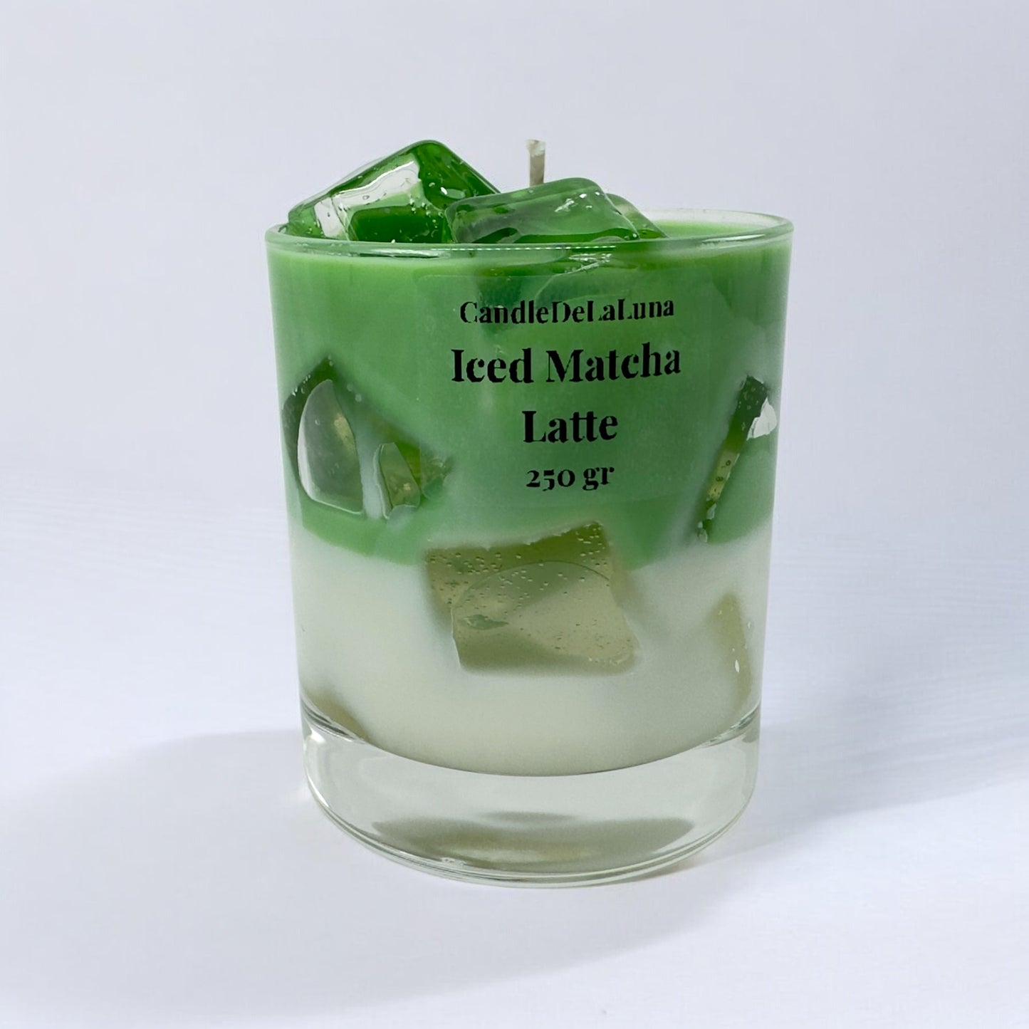 Eine handgemachte Kerze im Design von Iced Matcha Latte in einem niedrigen Glas mit grün-weißen Wachsschichten, künstlichen grünen Eiswürfeln auf der Oberseite und der Aufschrift „CandleDeLuna Iced Matcha Latte 250 gr“ vor einem schlichten weißen Hintergrund.
