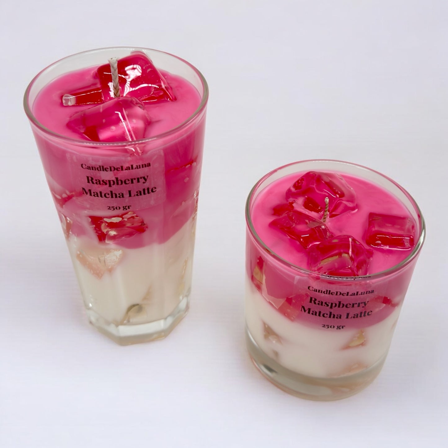 Zwei handgemachte Kerzen im Design von Raspberry Matcha Latte in einem hohen und einem niedrigen Glas mit rosa-weißen Wachsschichten, künstlichen rosa Eiswürfeln aus Wachs und der Aufschrift „CandleDeLaLuna Raspberry Matcha Latte 250 gr“ vor einem weißen Hintergrund.