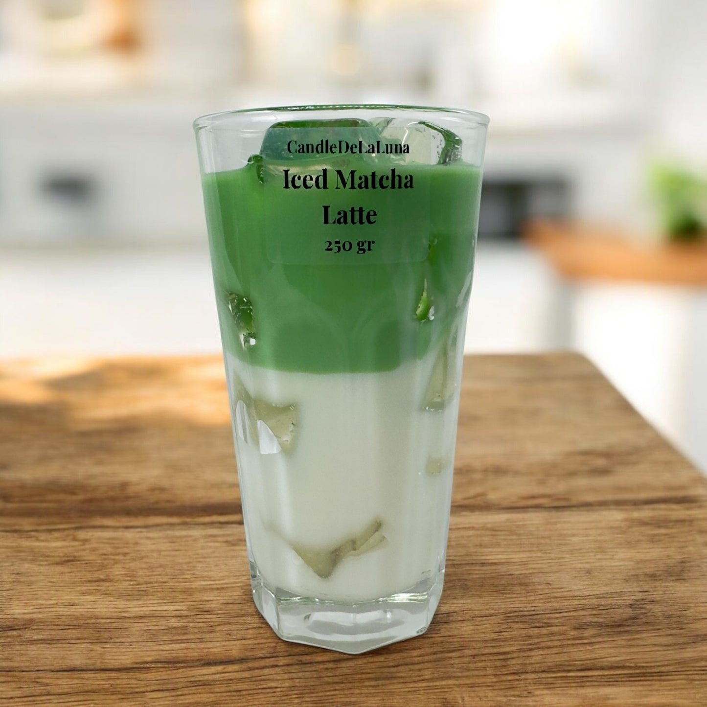 Eine handgemachte Kerze im Design von Iced Matcha Latte in einem hohen, facettierten Glas mit grün-weißen Wachsschichten, künstlichen grünen Eiswürfeln auf der Oberseite und der Aufschrift „CandleDeLuna Iced Matcha Latte 250 gr“, platziert auf einer Holzoberfläche vor einem verschwommenen Hintergrund.