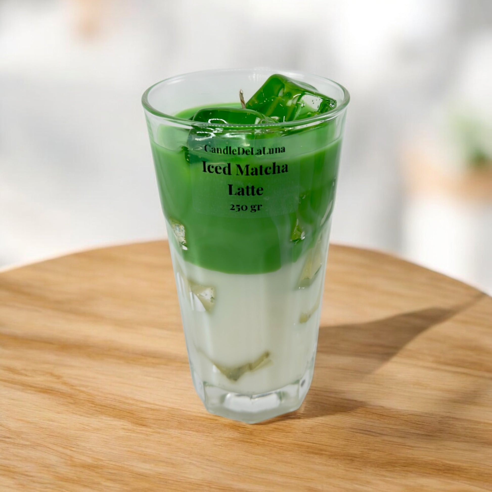 Eine handgemachte Kerze im Design von Iced Matcha Latte in einem hohen, facettierten Glas mit grün-weißen Wachsschichten, künstlichen grünen Eiswürfeln auf der Oberseite und der Aufschrift „CandleDeLuna Iced Matcha Latte 250 gr“, platziert auf einer runden Holzoberfläche im Sonnenlicht vor einem verschwommenen Hintergrund.