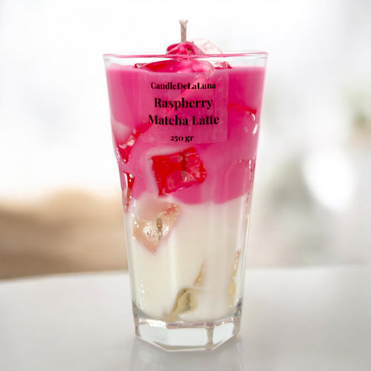 Eine dekorative Kerze in Form eines Raspberry Matcha Latte Getränks in einem Glas, bestehend aus einer weißen und einer leuchtend pinken Wachsschicht mit simulierten Eiswürfeln und einem Docht an der Spitze.