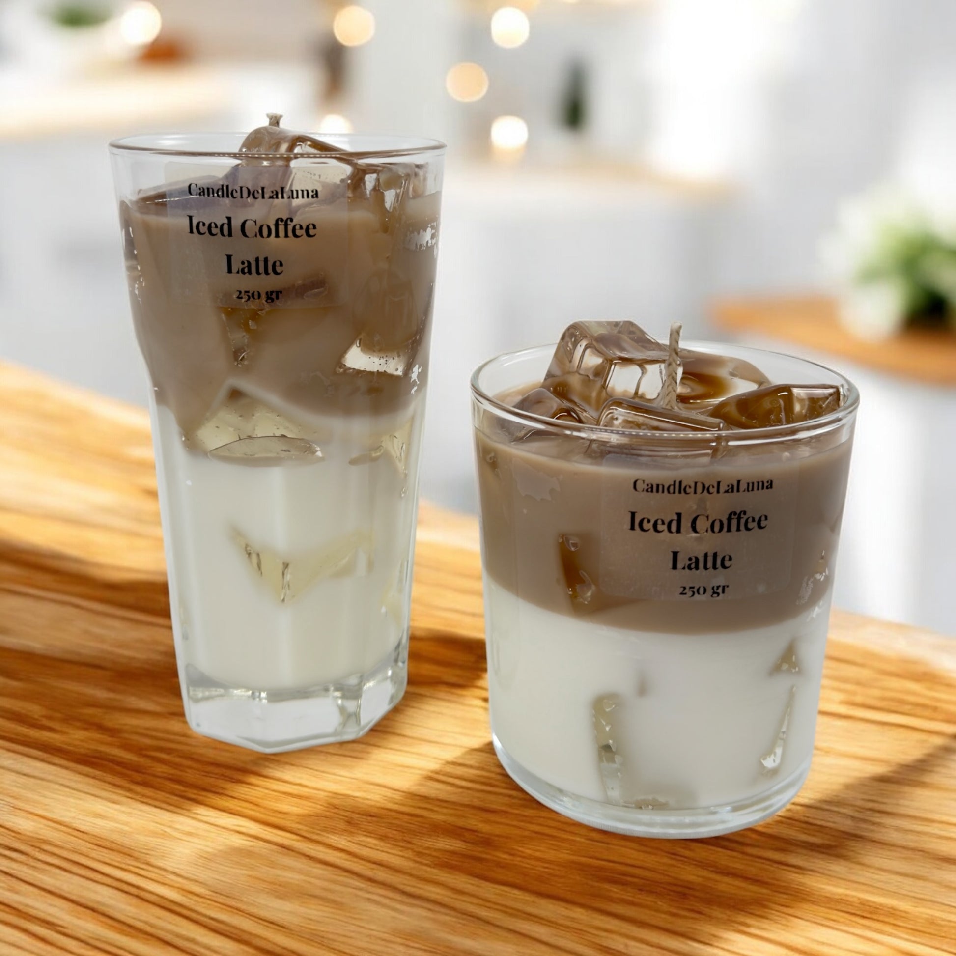 Zwei dekorative Kerzen in einem hohen und einem niedrigen Glas auf einer Holzoberfläche, gestaltet wie ein Eiskaffee-Latte mit einer weißen Schicht unten, einer hellbraunen Kaffeeschicht oben und transparenten Wachs-Eiswürfeln.
