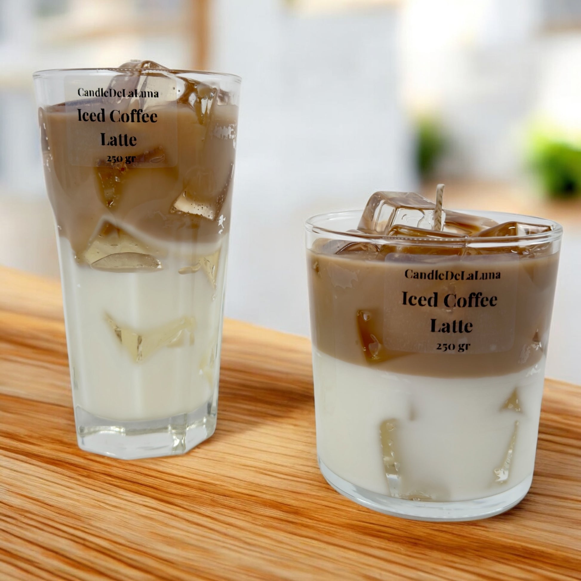 Zwei dekorative Kerzen in einem hohen und einem niedrigen Glas auf einer Holzoberfläche, gestaltet wie ein Eiskaffee-Latte mit einer weißen Schicht unten, einer hellbraunen Kaffeeschicht oben und transparenten Wachs-Eiswürfeln.
