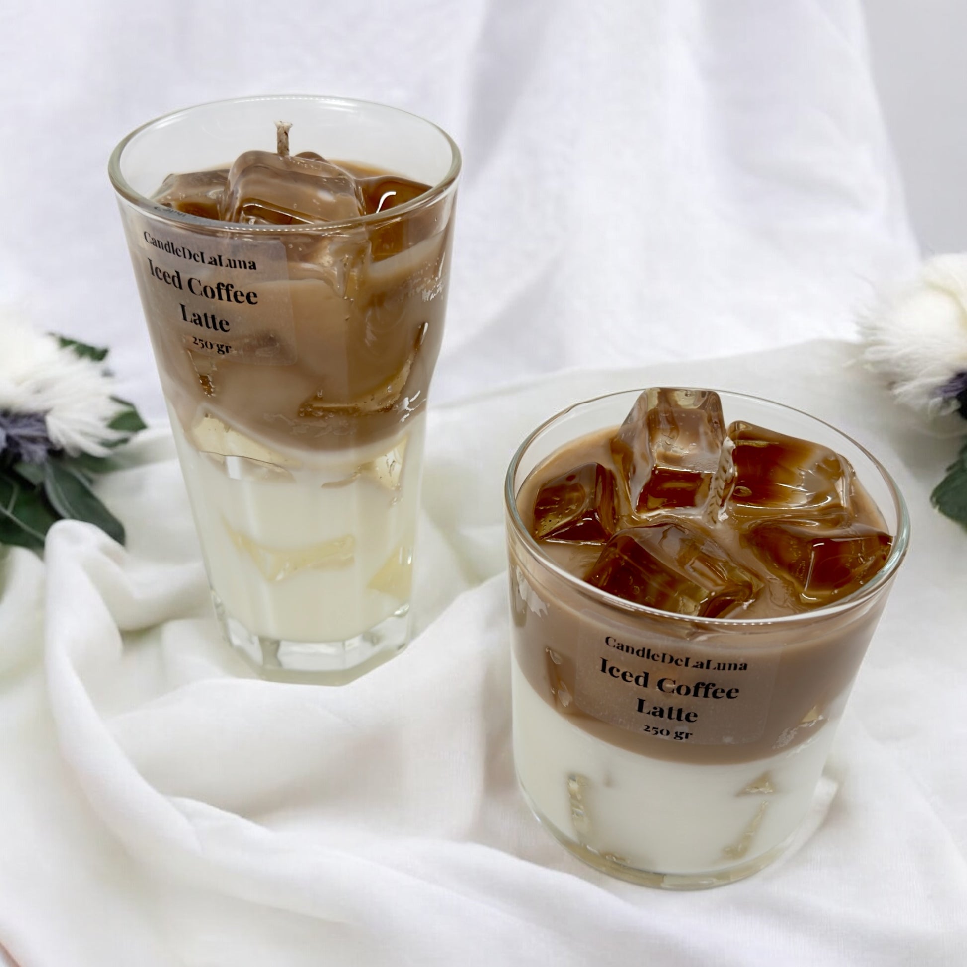 Zwei handgemachte Kerzen im Design von Iced Coffee Latte in einem hohen und einem niedrigen Glas mit braun-weißen Wachsschichten, künstlichen braunen Eiswürfeln auf der Oberseite und der Aufschrift „CandleDeLaLuna Iced Coffee Latte 250 gr“ vor einem weißen Stoffhintergrund.