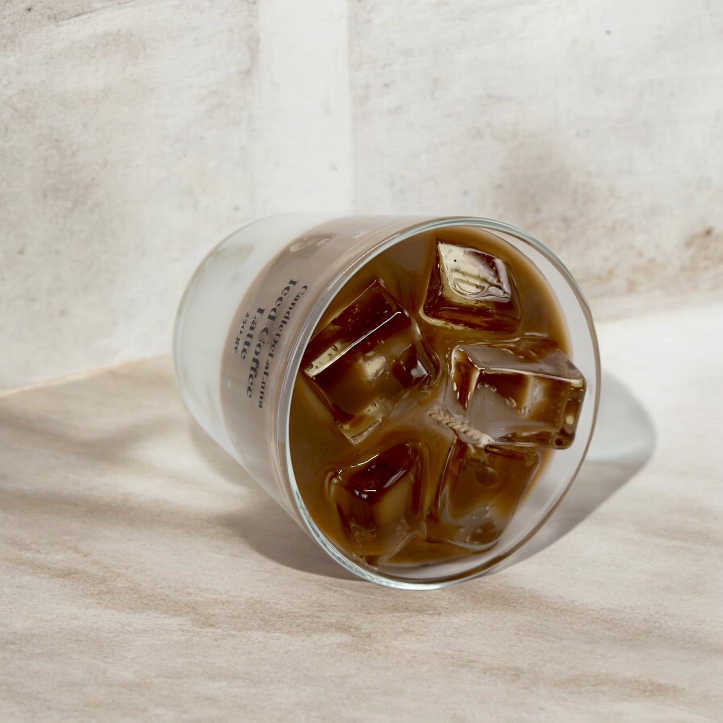 Eine liegende dekorative Kerze im Glas, gestaltet wie ein Eiskaffee-Latte mit einer hellbraunen Kaffeeschicht und realistisch wirkenden Wachs-Eiswürfeln an der Oberfläche.