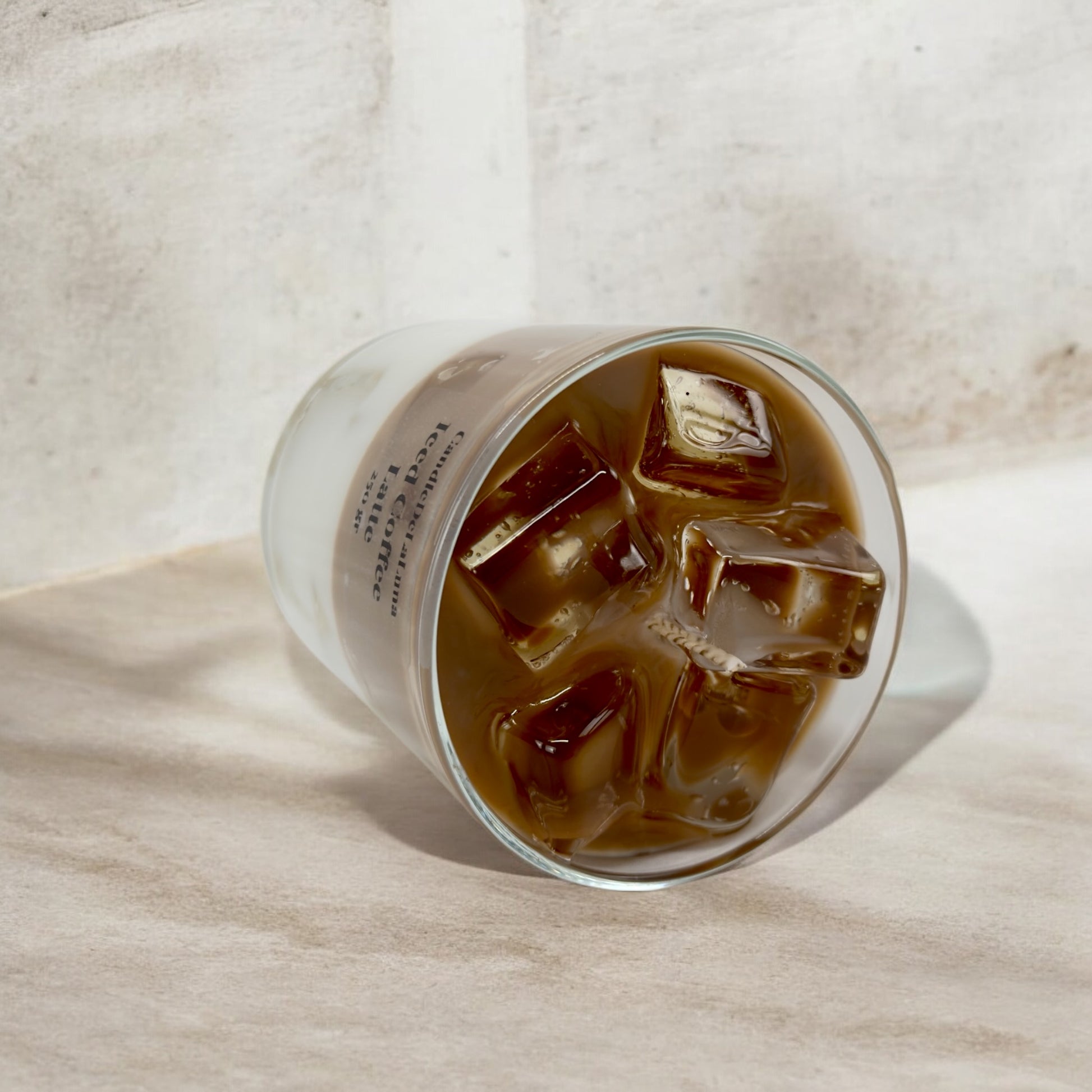 Eine liegende dekorative Kerze im Glas, gestaltet wie ein Eiskaffee-Latte mit einer hellbraunen Kaffeeschicht und realistisch wirkenden Wachs-Eiswürfeln an der Oberfläche.