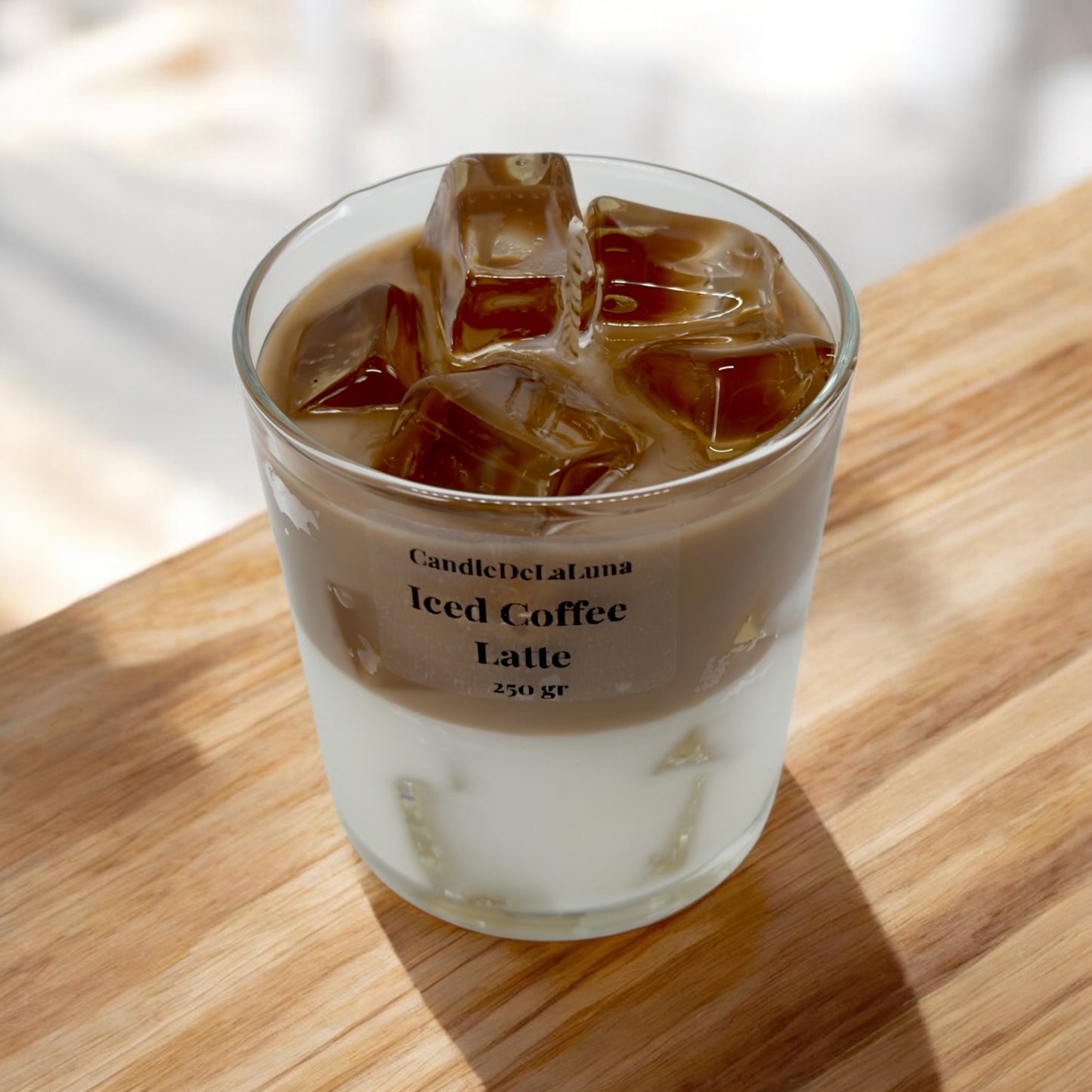Eine dekorative Kerze im Glas auf einer Holzoberfläche, gestaltet wie ein Eiskaffee-Latte mit einer weißen Schicht unten, einer hellbraunen Kaffeeschicht oben und realistisch wirkenden Wachs-Eiswürfeln an der Oberfläche.