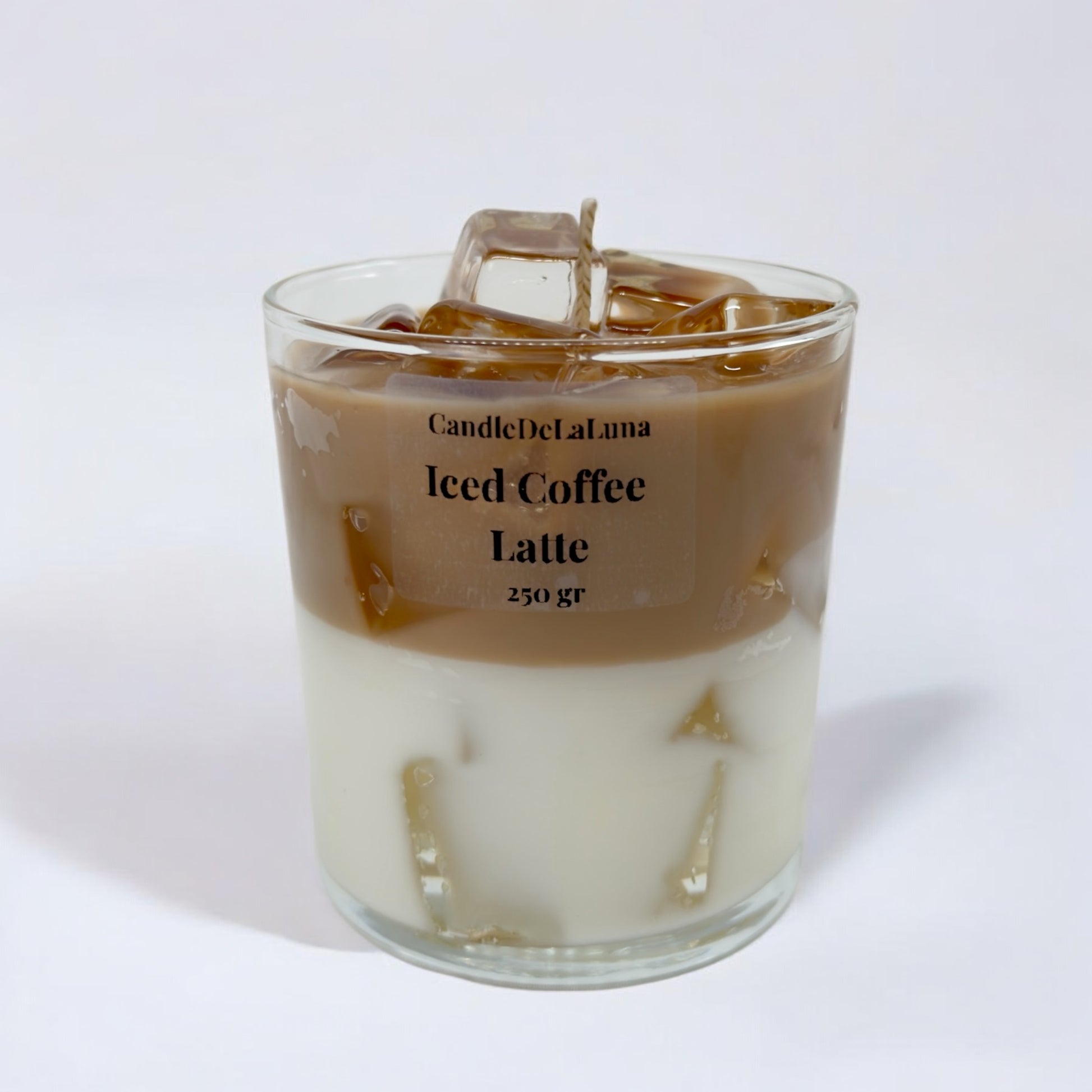 Eine dekorative Kerze im Glas auf weißem Hintergrund, gestaltet wie ein Eiskaffee-Latte mit einer weißen Schicht unten, einer hellbraunen Kaffeeschicht oben und transparenten Wachs-Eiswürfeln.