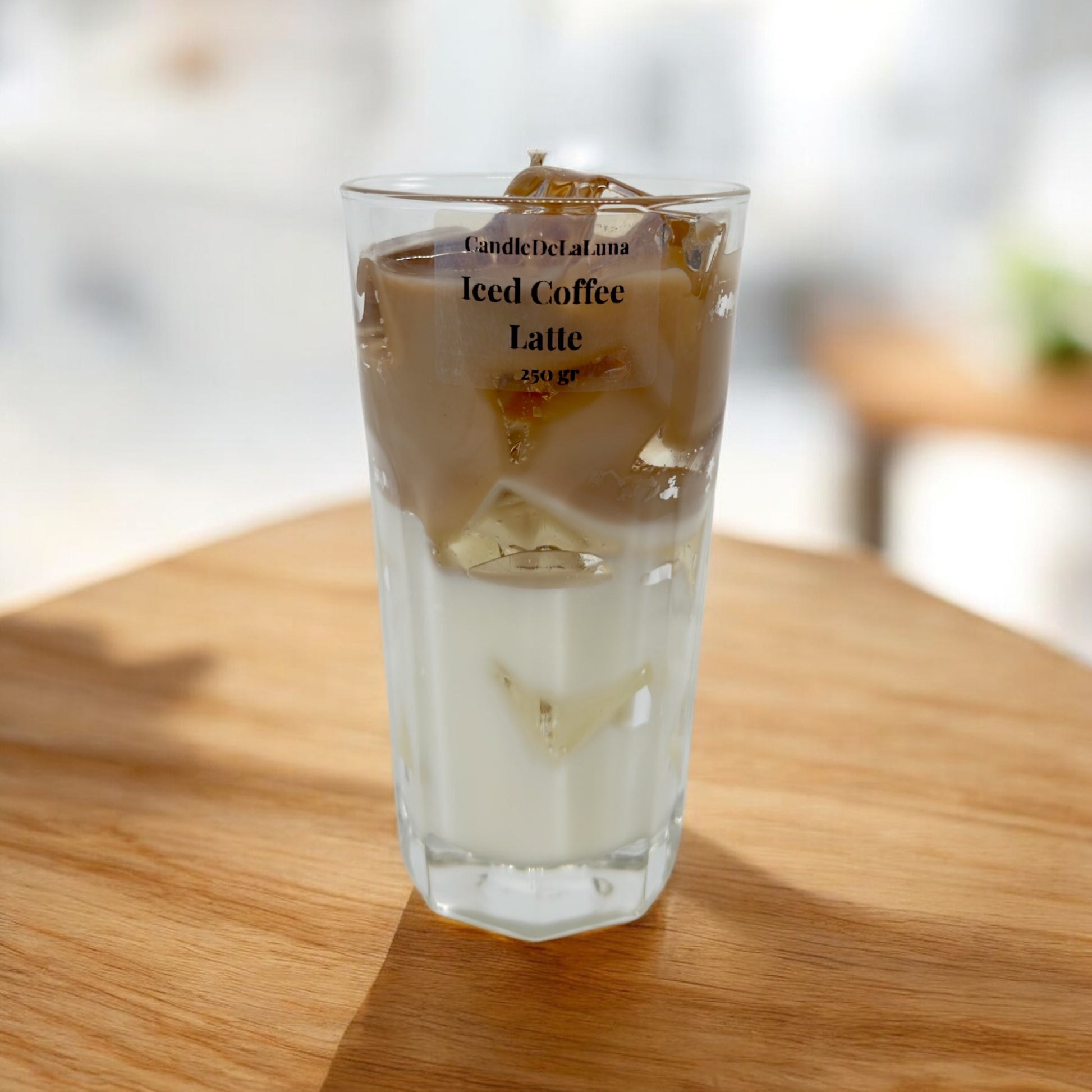 Eine dekorative Kerze im hohen Glas auf einer Holzoberfläche, gestaltet wie ein Eiskaffee-Latte mit einer weißen Schicht unten, einer hellbraunen Kaffeeschicht oben und transparenten Wachs-Eiswürfeln.