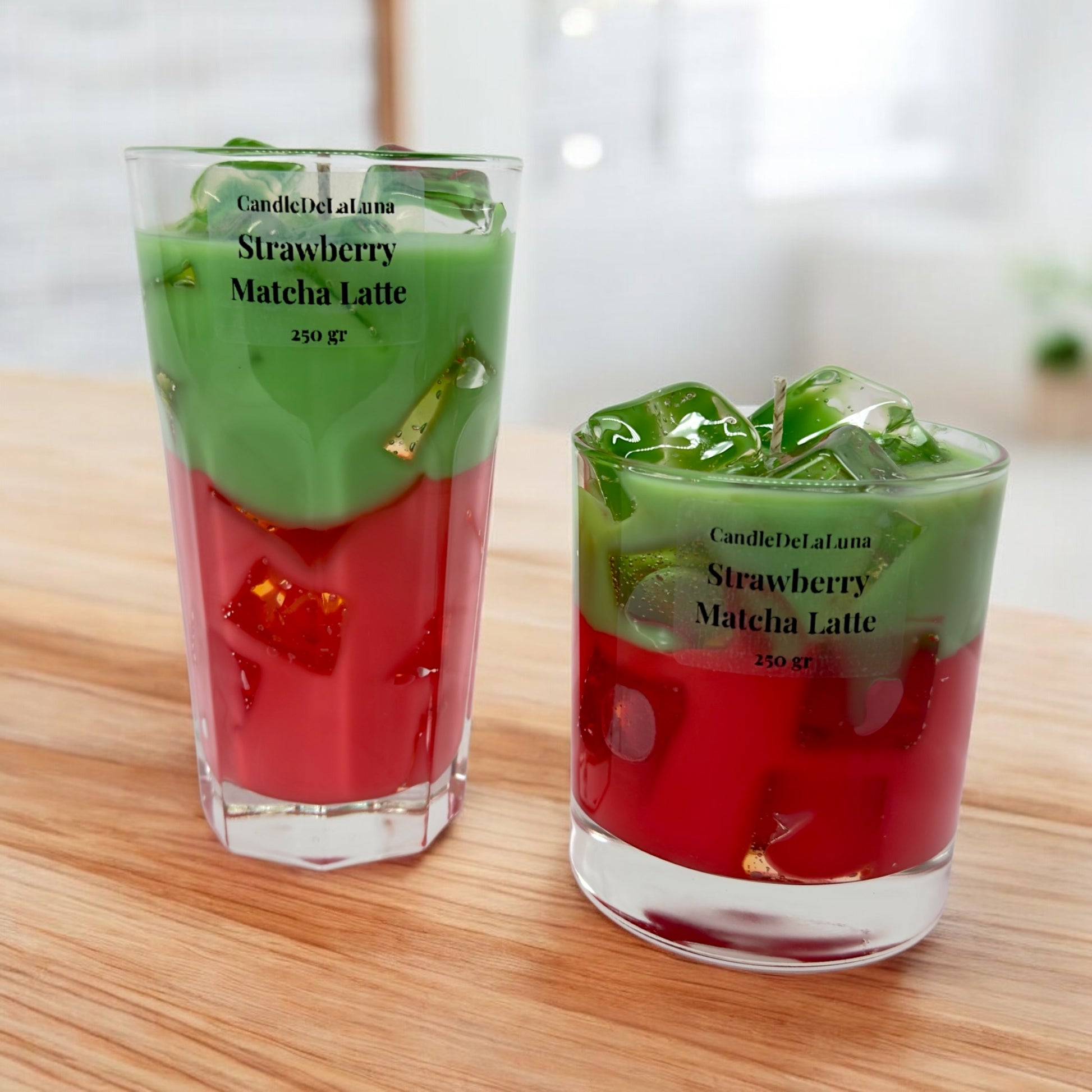 Zwei dekorative Kerzen in Gläsern unterschiedlicher Größe, gestaltet wie ein eisgekühlter Strawberry Matcha Latte, mit einer roten Schicht unten, einer grünen Schicht oben und transparenten Wachs-Eiswürfeln auf der Oberfläche.