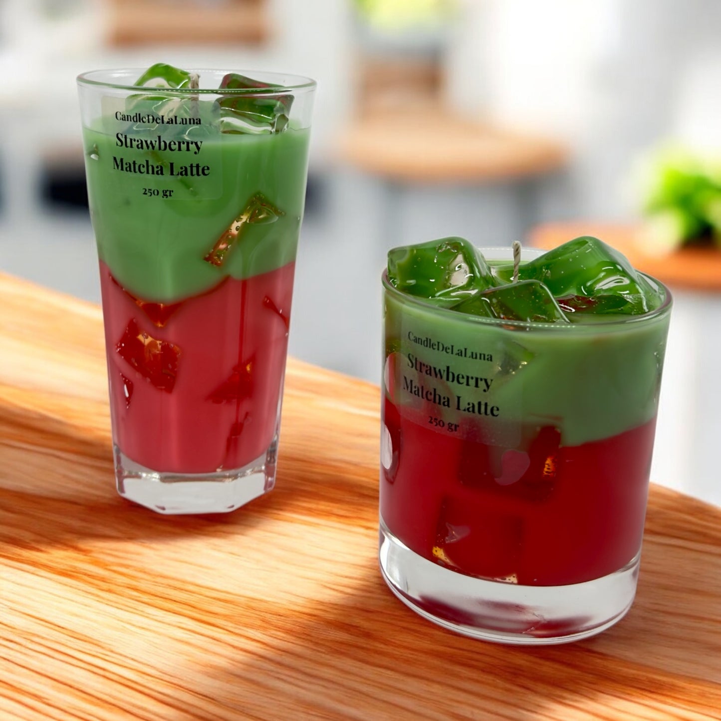 Eine dekorative Kerze im hohen Glas und eine im niedrigen Glas, gestaltet wie ein eisgekühlter Strawberry Matcha Latte mit einer roten Schicht unten, einer grünen Schicht oben und transparenten Wachs-Eiswürfeln.