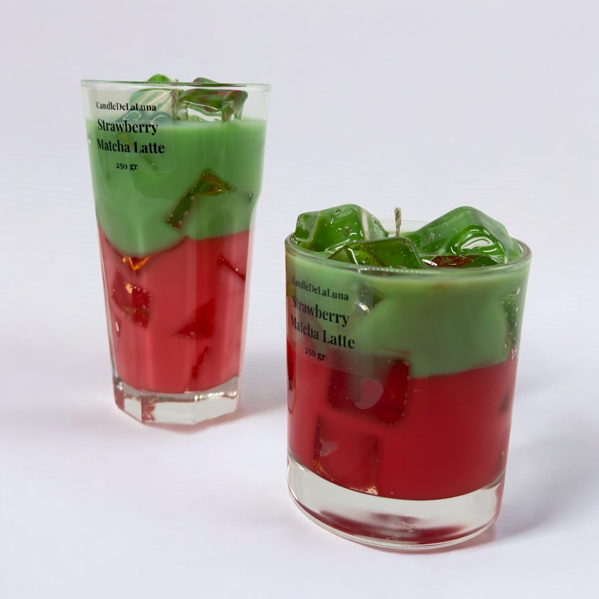 Eine dekorative Kerze im hohen Glas und eine im niedrigen Glas auf weißem Hintergrund, gestaltet wie ein eisgekühlter Strawberry Matcha Latte mit einer roten Schicht unten, einer grünen Schicht oben und transparenten Wachs-Eiswürfeln.