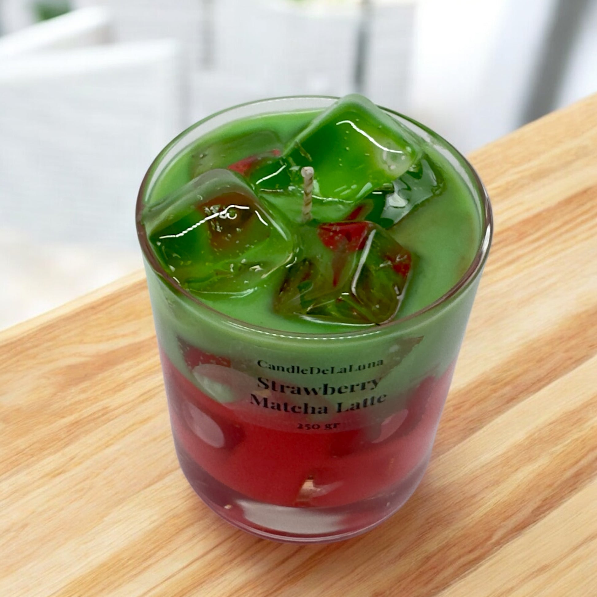 Eine dekorative Kerze im Glas, die wie ein eisgekühlter Strawberry Matcha Latte gestaltet ist, mit einer roten Schicht unten, einer grünen Matcha-Schicht oben und realistisch wirkenden Wachs-Eiswürfeln an der Oberfläche.