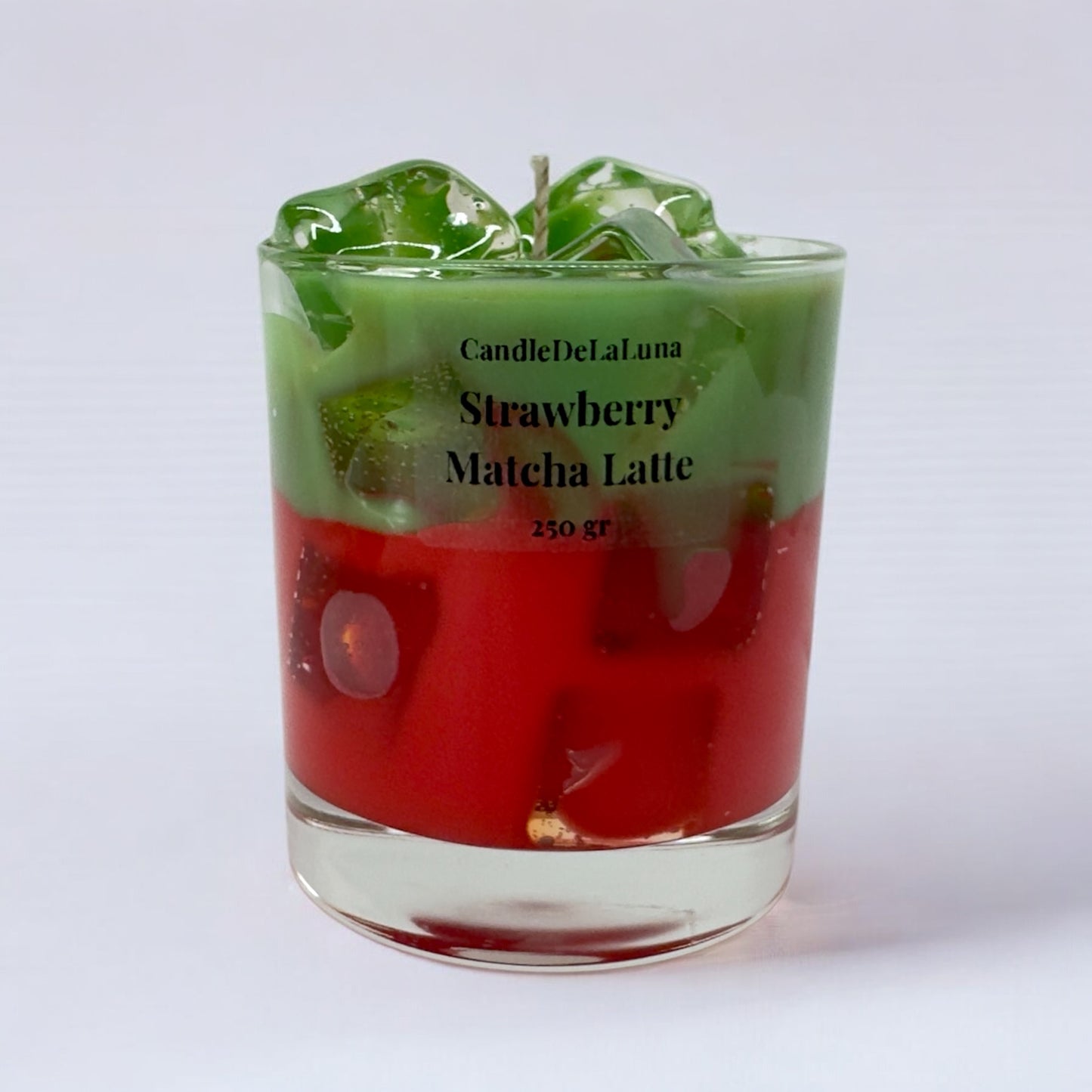 Eine dekorative Kerze im niedrigen Glas auf weißem Hintergrund, gestaltet wie ein eisgekühlter Strawberry Matcha Latte mit einer roten Schicht unten, einer grünen Schicht oben und transparenten Wachs-Eiswürfeln.