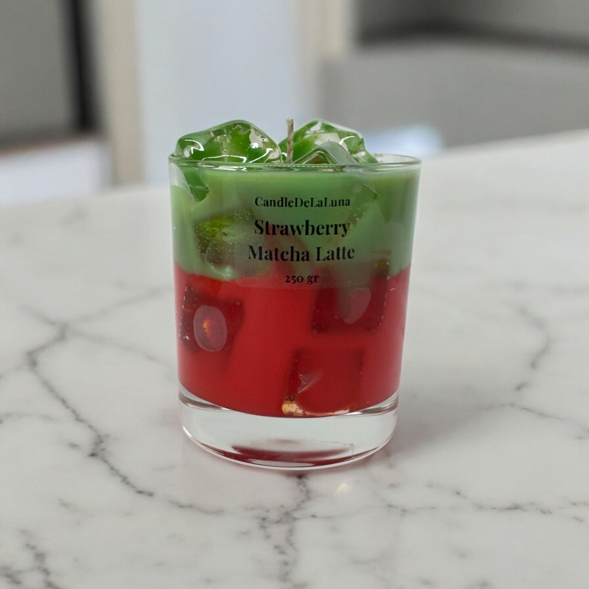 Eine dekorative Kerze im niedrigen Glas auf einer hellen Marmoroberfläche, gestaltet wie ein eisgekühlter Strawberry Matcha Latte mit einer roten Schicht unten, einer grünen Schicht oben und transparenten Wachs-Eiswürfeln.