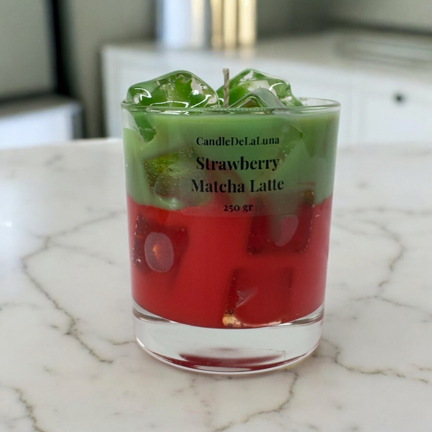Eine dekorative Kerze im niedrigen Glas, gestaltet wie ein eisgekühlter Strawberry Matcha Latte mit einer roten Schicht unten, einer grünen Schicht oben und transparenten Wachs-Eiswürfeln.