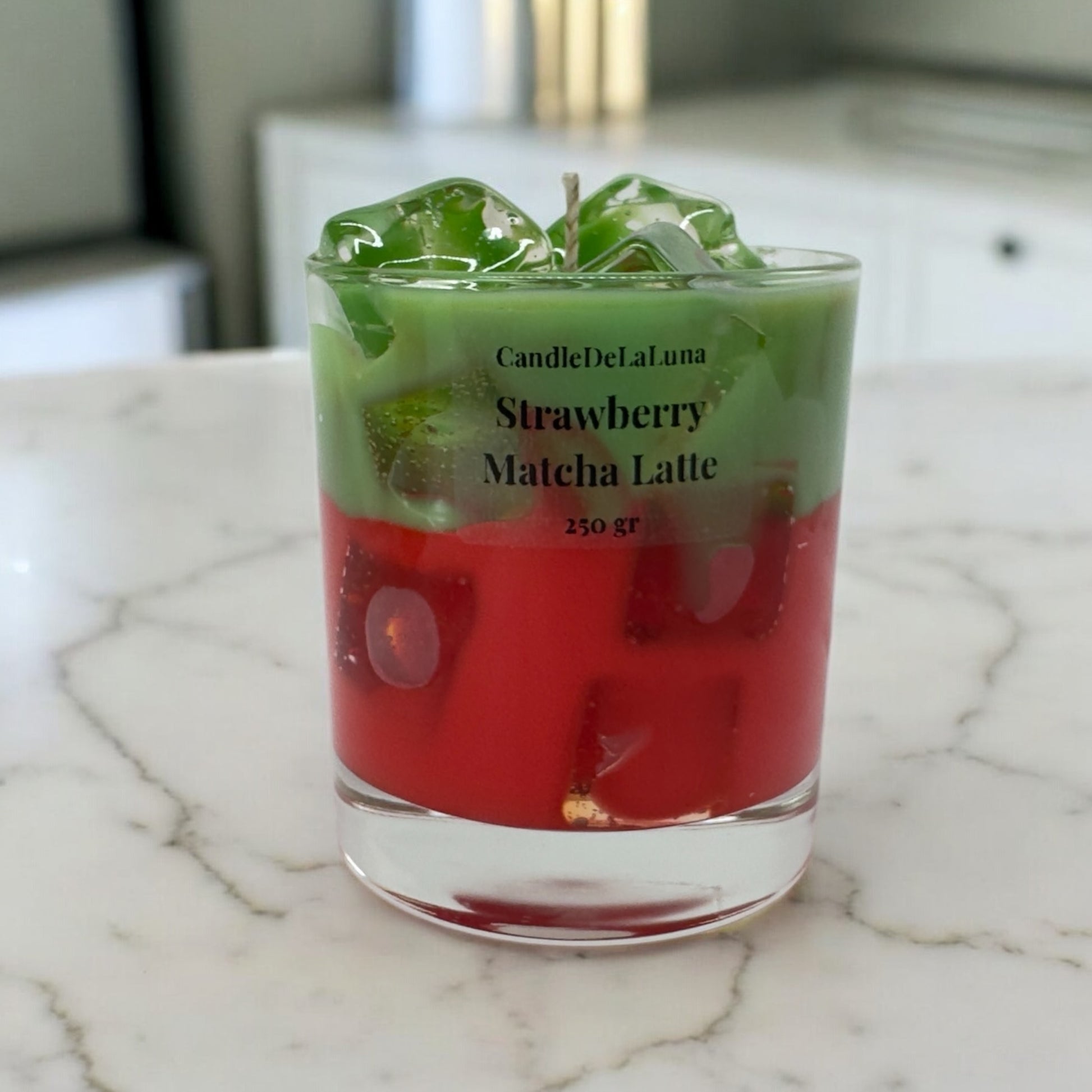 Eine dekorative Kerze im niedrigen Glas, gestaltet wie ein eisgekühlter Strawberry Matcha Latte mit einer roten Schicht unten, einer grünen Schicht oben und transparenten Wachs-Eiswürfeln.