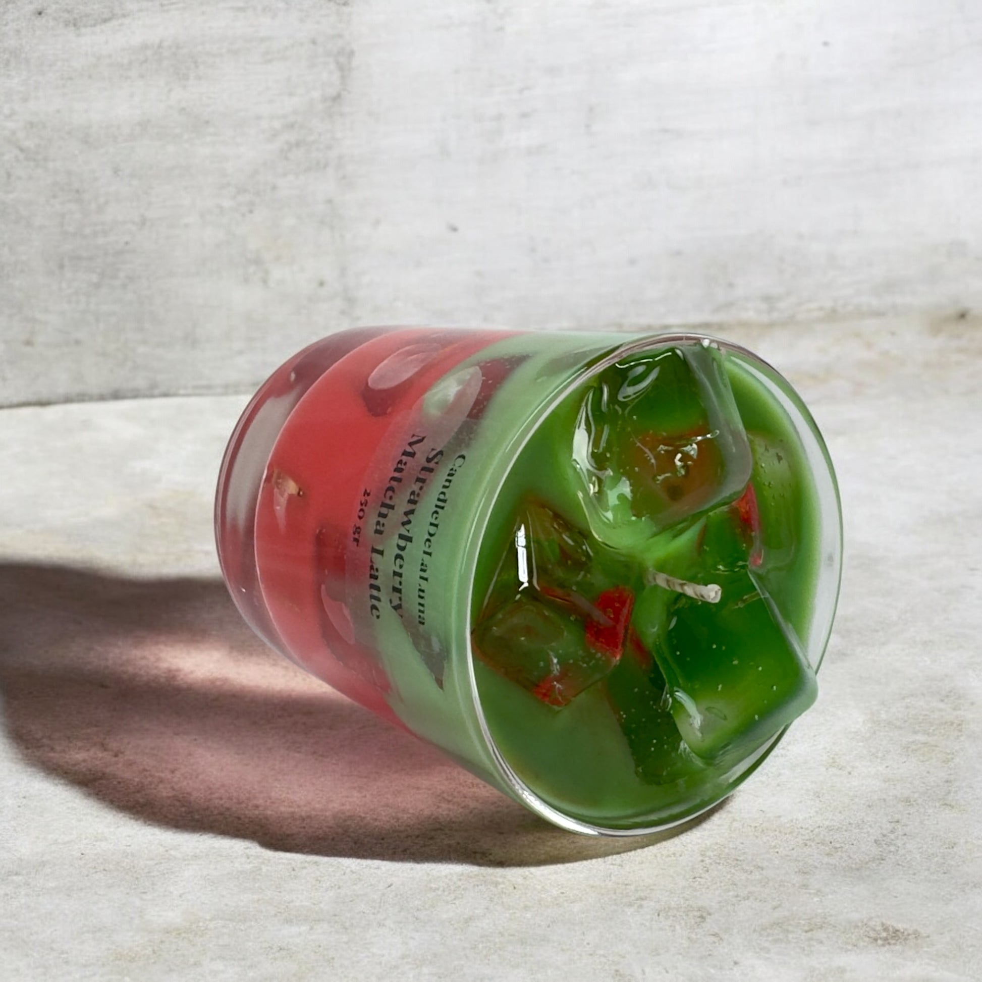 Eine liegende dekorative Kerze im Glas, gestaltet wie ein eisgekühlter Strawberry Matcha Latte mit einer roten und einer grünen Schicht sowie Wachs-Eiswürfeln auf der Oberseite.