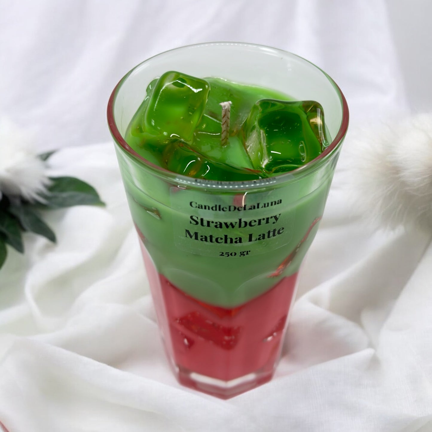 Eine dekorative Kerze im hohen Glas auf weißem Stoffhintergrund, gestaltet wie ein eisgekühlter Strawberry Matcha Latte mit einer roten Schicht unten, einer grünen Schicht oben und transparenten Wachs-Eiswürfeln.