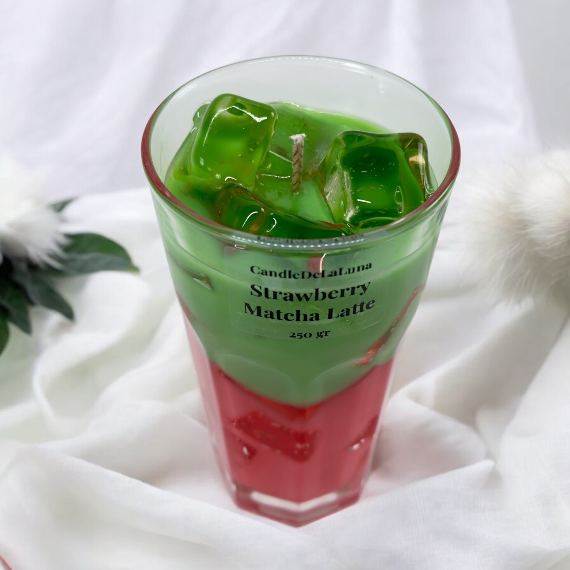 Eine dekorative Kerze im hohen Glas auf weißem Stoffhintergrund, gestaltet wie ein eisgekühlter Strawberry Matcha Latte mit einer roten Schicht unten, einer grünen Schicht oben und transparenten Wachs-Eiswürfeln.