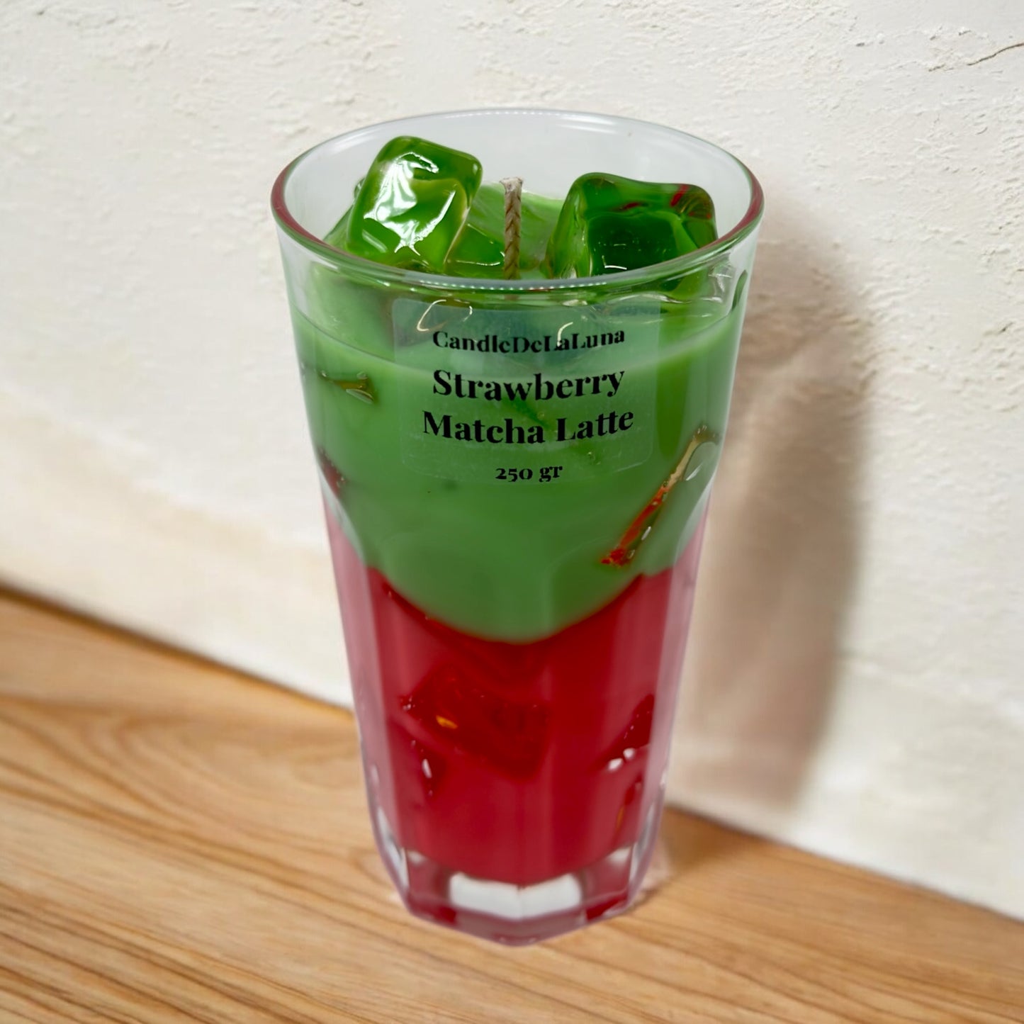 Eine dekorative Kerze im hohen Glas auf einer Holzoberfläche vor einer weißen Wand, gestaltet wie ein eisgekühlter Strawberry Matcha Latte mit einer roten Schicht unten, einer grünen Schicht oben und transparenten Wachs-Eiswürfeln.