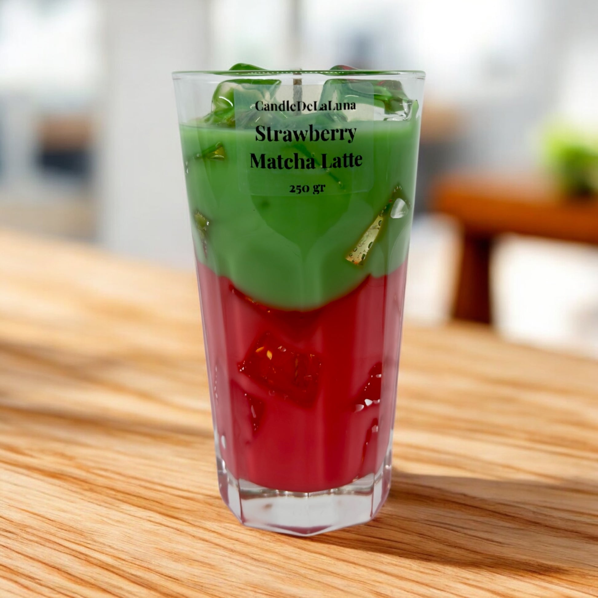 Eine dekorative Kerze im hohen Glas, gestaltet wie ein eisgekühlter Strawberry Matcha Latte, mit einer roten Schicht unten, einer grünen Matcha-Schicht oben und Wachs-Eiswürfeln an der Oberfläche.