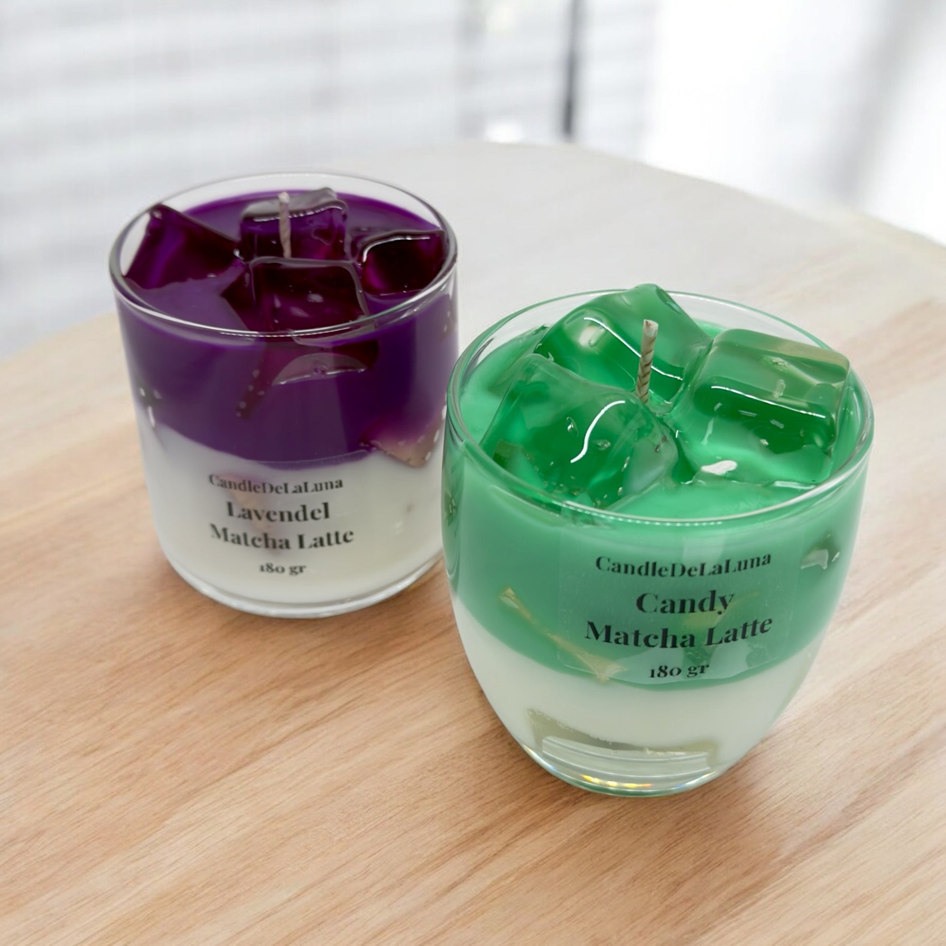 Zwei dekorative Kerzen in Gläsern auf einer Holzoberfläche, eine gestaltet als lila Lavendel Matcha Latte und die andere als grüner Candy Matcha Latte, beide mit einer weißen Unterschicht und transparenten Wachs-Eiswürfeln.