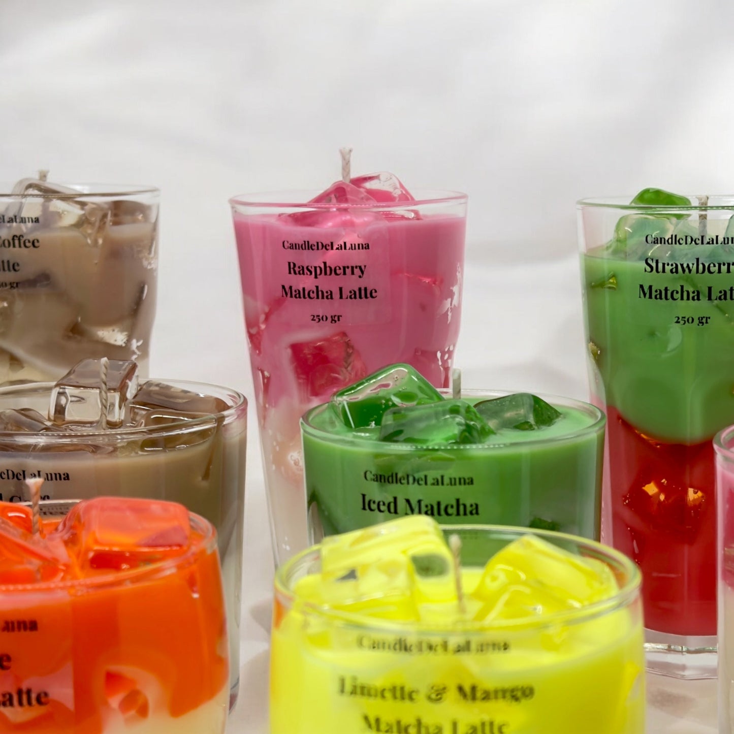 Eine Gruppe von verschiedenen handgemachten Kerzen im Design von Eiskaffee- und Matcha-Getränken in hohen und niedrigen Gläsern mit bunten Wachsschichten, künstlichen Eiswürfeln auf der Oberseite und Etiketten wie „Raspberry Matcha Latte“, „Strawberry Matcha Latte“, „Iced Matcha“ und „Limette & Mango Matcha Latte“ vor einem weißen Hintergrund.