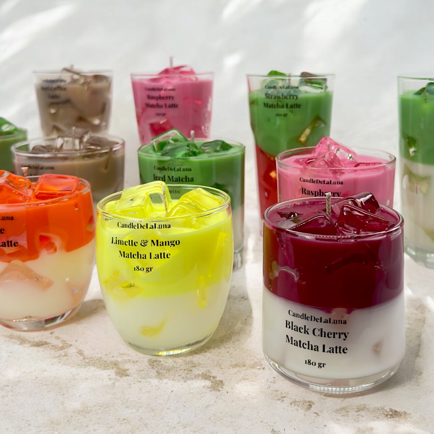 Eine Vielzahl von verschiedenen handgemachten Kerzen im Design von Kaltgetränken in hohen und niedrigen Gläsern mit bunten Wachsschichten, künstlichen Eiswürfeln auf der Oberseite und Etiketten wie „Black Cherry Matcha Latte“, „Limette & Mango Matcha Latte“, „Raspberry“ und „Strawberry Matcha Latte“ auf einer hellen Oberfläche vor einem hellen Hintergrund.