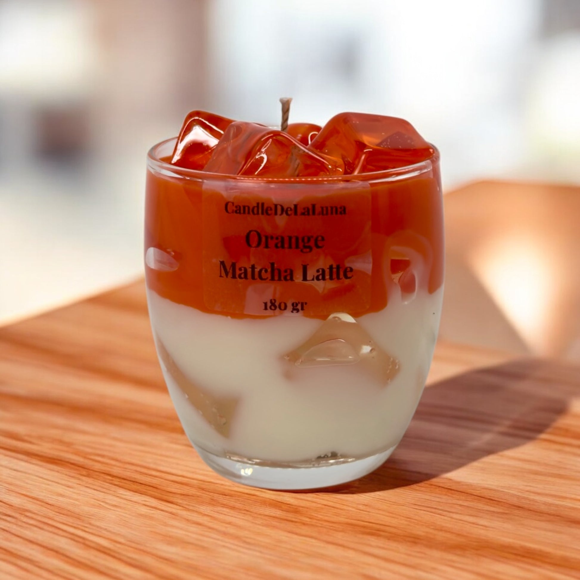 Eine handgemachte Kerze im Design von Orange Matcha Latte in einem niedrigen Glas mit orange-weißen Wachsschichten, künstlichen orangefarbenen Eiswürfeln auf der Oberseite und der Aufschrift „CandleDeLaLuna Orange Matcha Latte 180 gr“, platziert auf einer Holzoberfläche im Sonnenlicht vor einem verschwommenen Hintergrund.