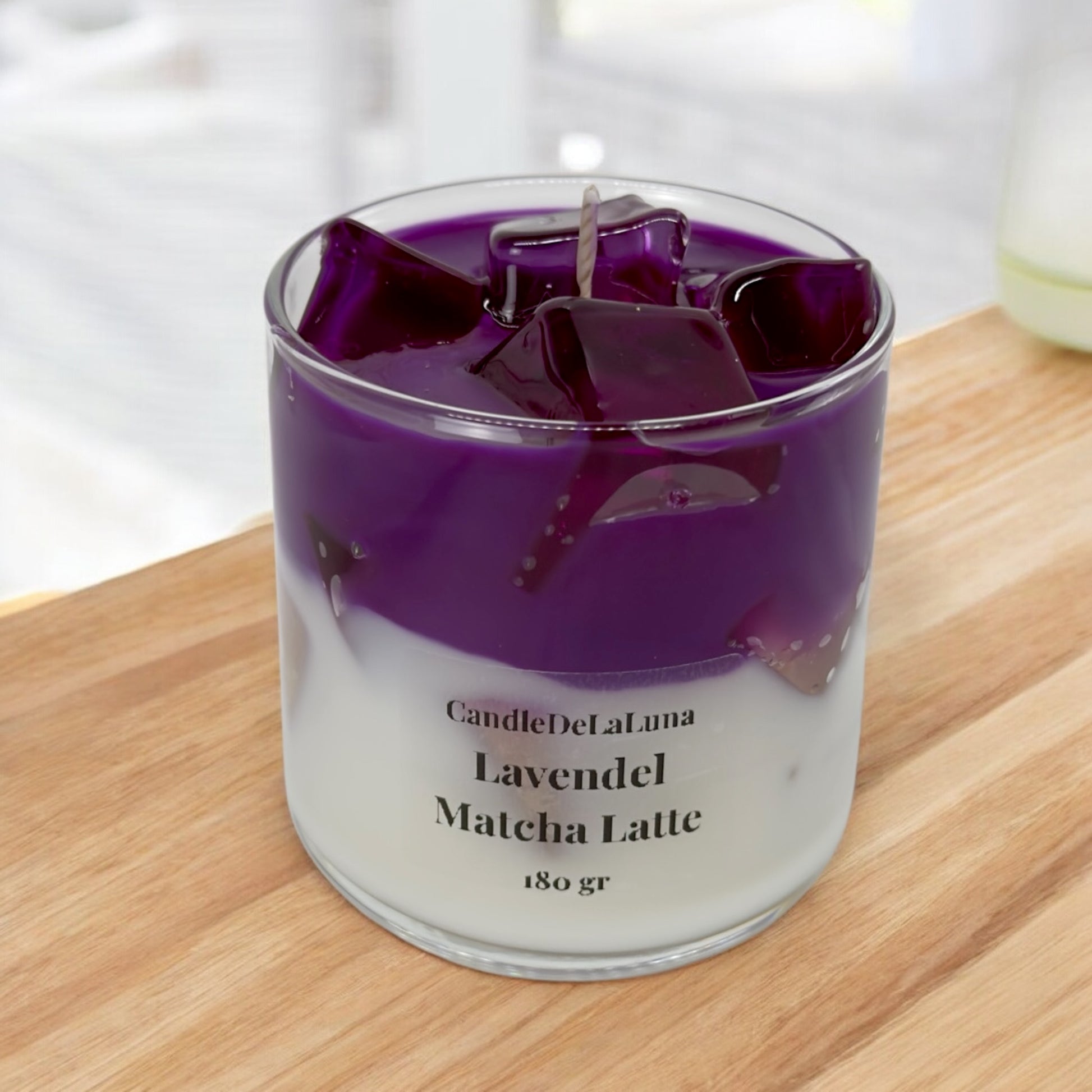 Eine handgemachte Kerze im Design von Lavendel Matcha Latte in einem niedrigen Glas mit violett-weißen Wachsschichten, künstlichen violetten Eiswürfeln auf der Oberseite und der Aufschrift „CandleDeLaLuna Lavendel Matcha Latte 180 gr“, platziert auf einer Holzoberfläche vor einem hellen Hintergrund.