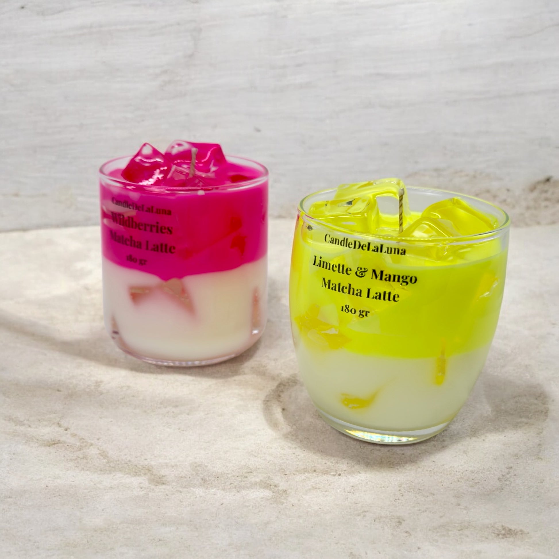 Zwei dekorative Kerzen in Gläsern, eine gestaltet als pinker Wildberries Matcha Latte und die andere als gelber Limette & Mango Matcha Latte, beide mit einer weißen Unterschicht und Wachs-Eiswürfeln.