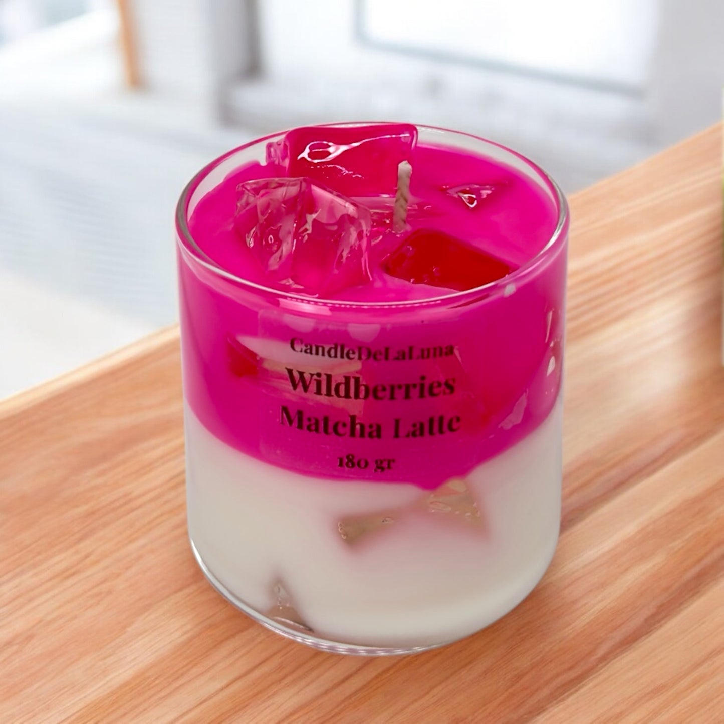 Eine dekorative Kerze im Glas, gestaltet wie ein Wildberries Matcha Latte mit einer weißen Schicht unten, einer leuchtend pinken Schicht oben und transparenten Wachs-Eiswürfeln an der Oberfläche.
