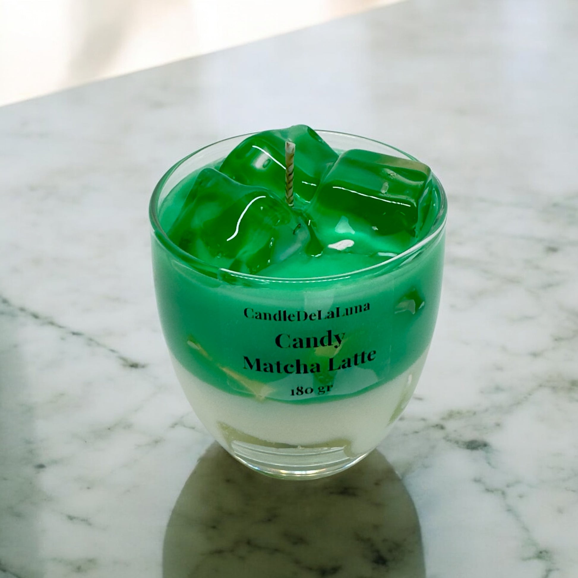 Eine dekorative Kerze im Glas auf einer Marmoroberfläche, gestaltet wie ein Candy Matcha Latte mit einer weißen Schicht unten, einer grünen Matcha-Schicht oben und transparenten Wachs-Eiswürfeln.