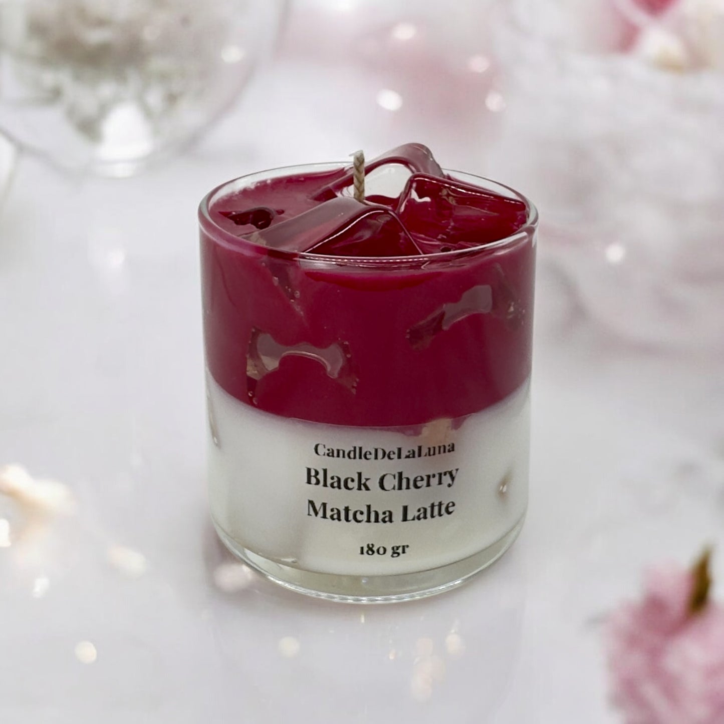 Eine dekorative Kerze im Glas, gestaltet wie ein Black Cherry Matcha Latte mit einer weißen Schicht unten, einer dunkelroten Kirschschicht oben und transparenten Wachs-Eiswürfeln an der Oberfläche.