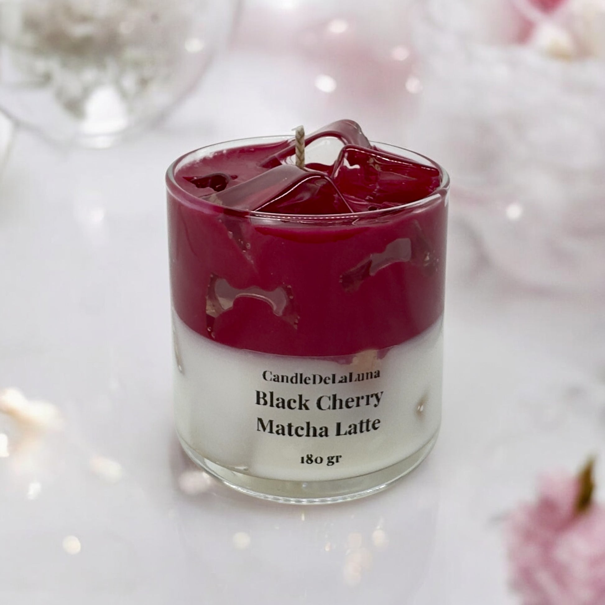 Eine dekorative Kerze im Glas, gestaltet wie ein Black Cherry Matcha Latte mit einer weißen Schicht unten, einer dunkelroten Kirschschicht oben und transparenten Wachs-Eiswürfeln an der Oberfläche.