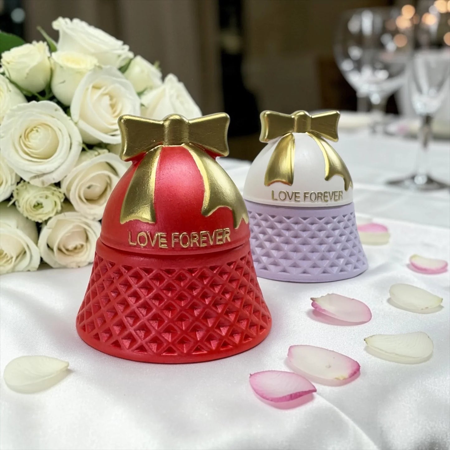 Rote und lavendelfarbene „Love Forever“ Kerzen festlich dekoriert mit weißen Rosen und Blütenblättern auf einer weißen Tischdecke im Restaurant.
