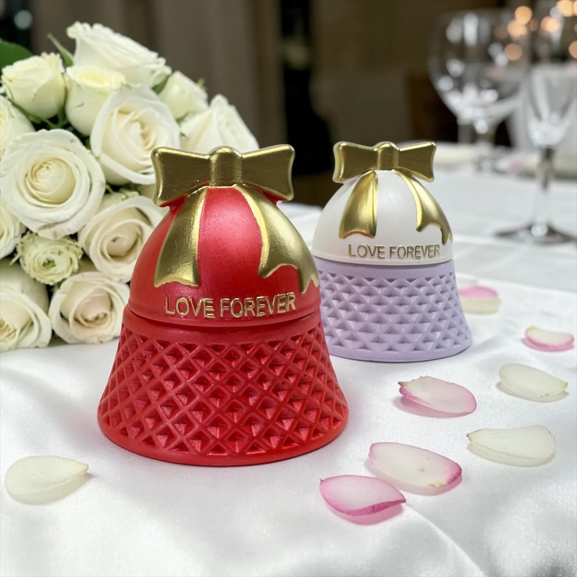 Rote und lavendelfarbene „Love Forever“ Kerzen festlich dekoriert mit weißen Rosen und Blütenblättern auf einer weißen Tischdecke im Restaurant.
