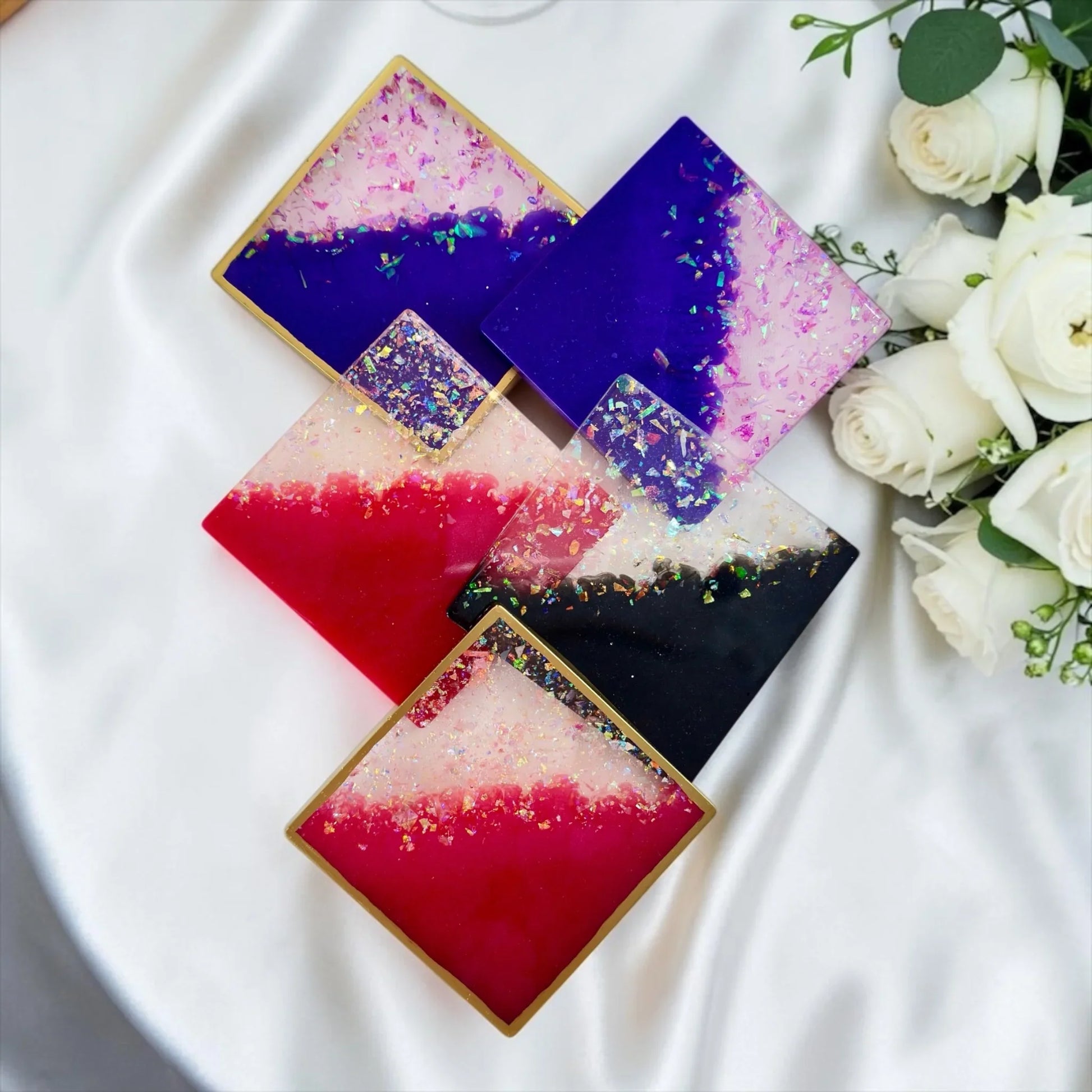 Fünf quadratische Epoxidharz-Untersetzer in Lila, Pink und Schwarz mit Goldrand, dekorativ arrangiert auf weißem Satin neben weißen Rosen.
