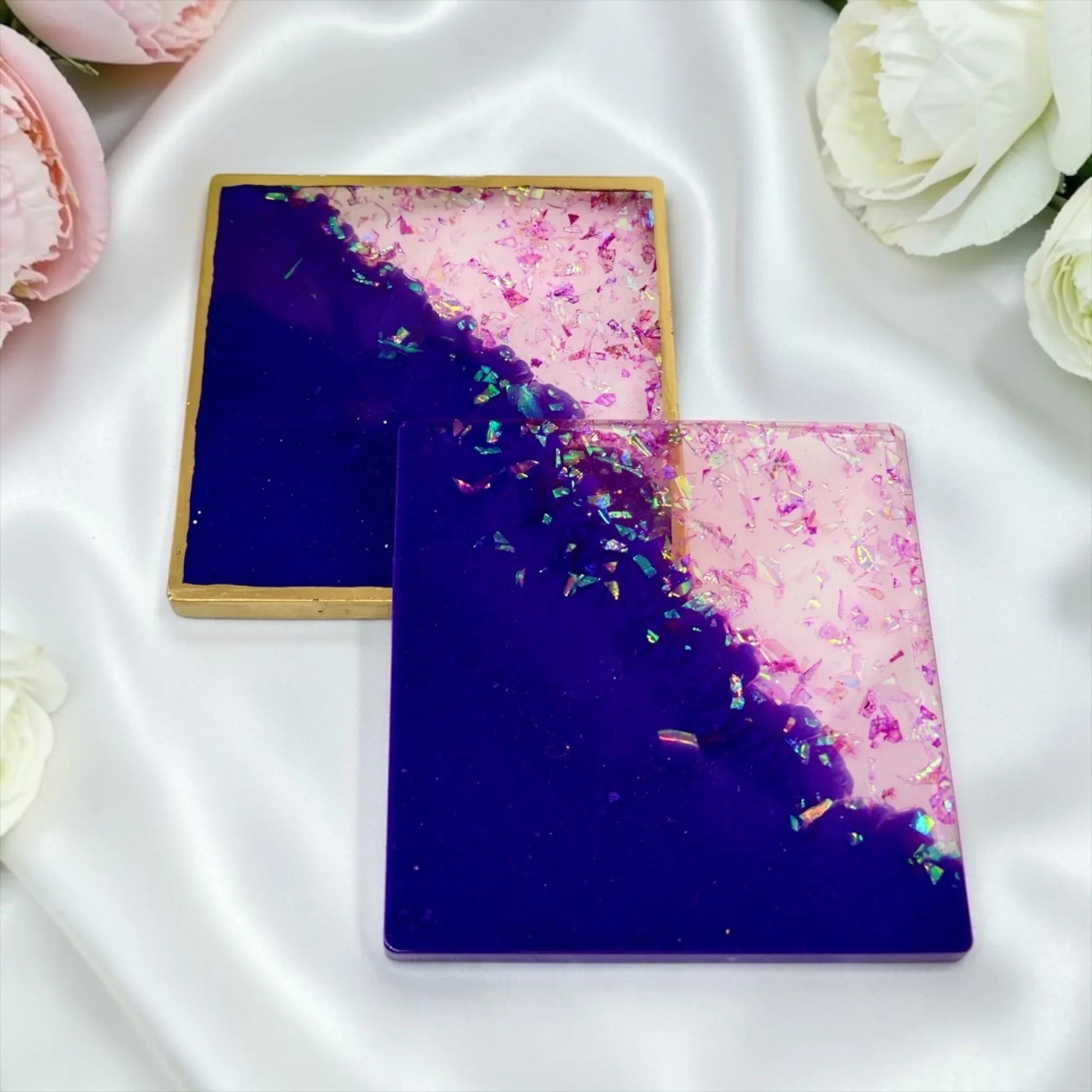 Zwei quadratische Untersetzer aus Epoxidharz in Dunkellila und Rosa mit Glitzer-Akzenten, einer davon mit Goldrand, auf weißem Satin neben hellen Rosen.
