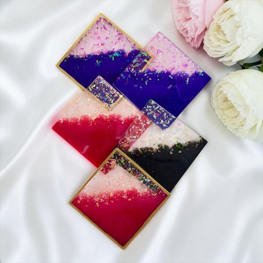 Fünf handgefertigte Untersetzer aus Epoxidharz in Lila, Pink und Schwarz mit Goldkanten, dekorativ platziert auf weißem Satin neben hellrosa Pfingstrosen.

