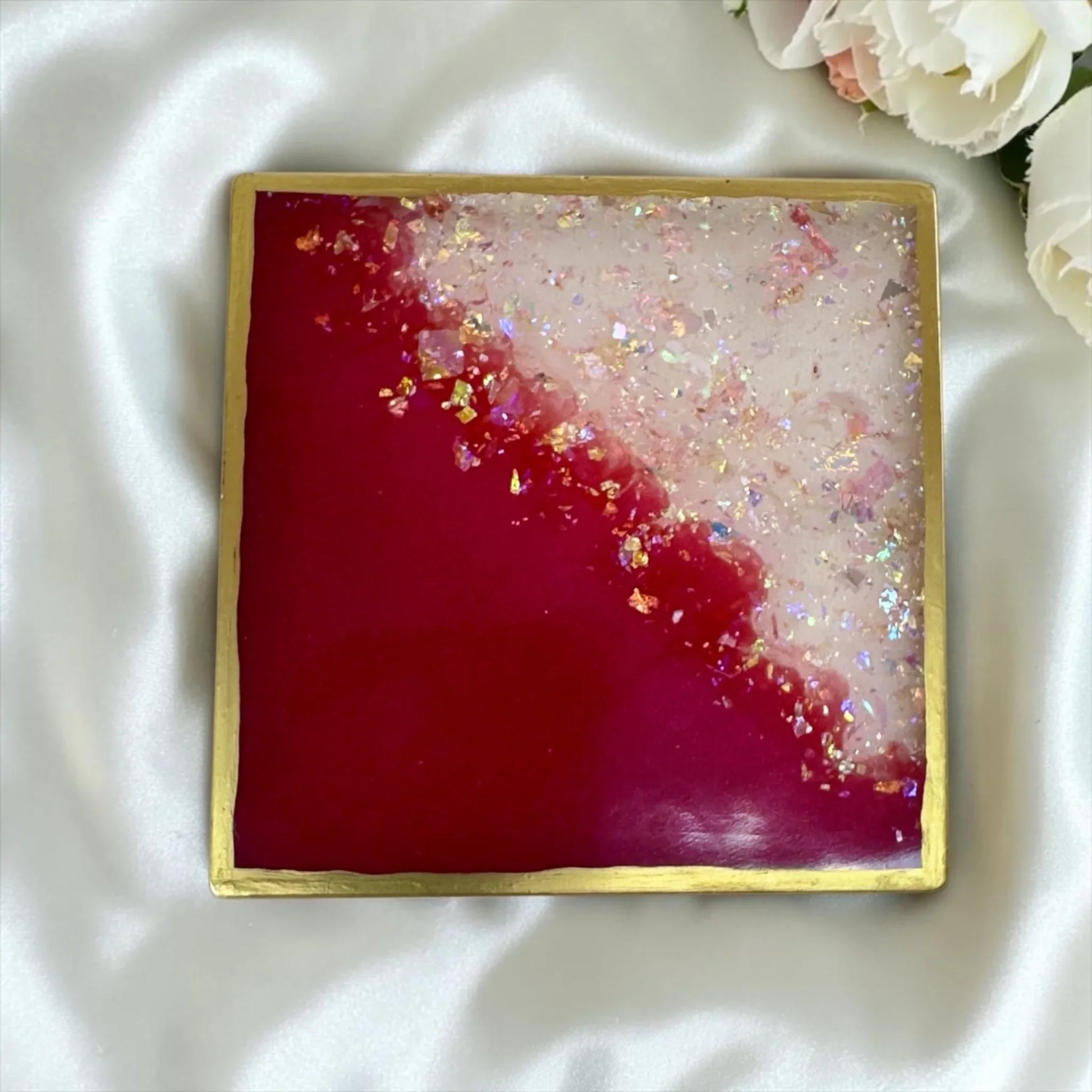 Ein quadratischer Untersetzer aus Epoxidharz in Pink und Weiß mit Goldrand и Glitzerflocken auf weißem Satin.