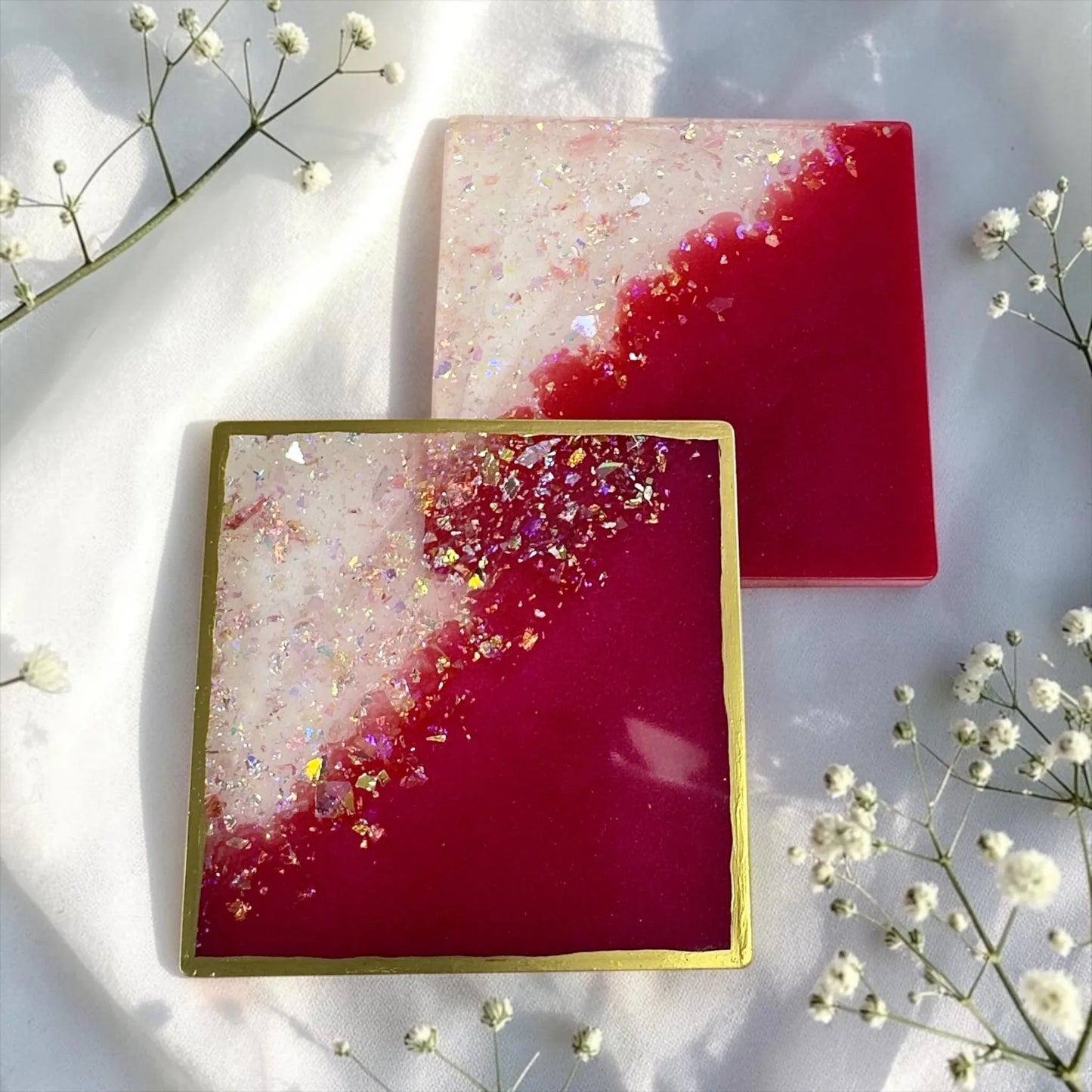 Zwei quadratische Epoxidharz-Untersetzer in Pink und Weiß mit Glitzer, einer davon mit Goldrand, umgeben von weißen Schleierkraut-Blumen.