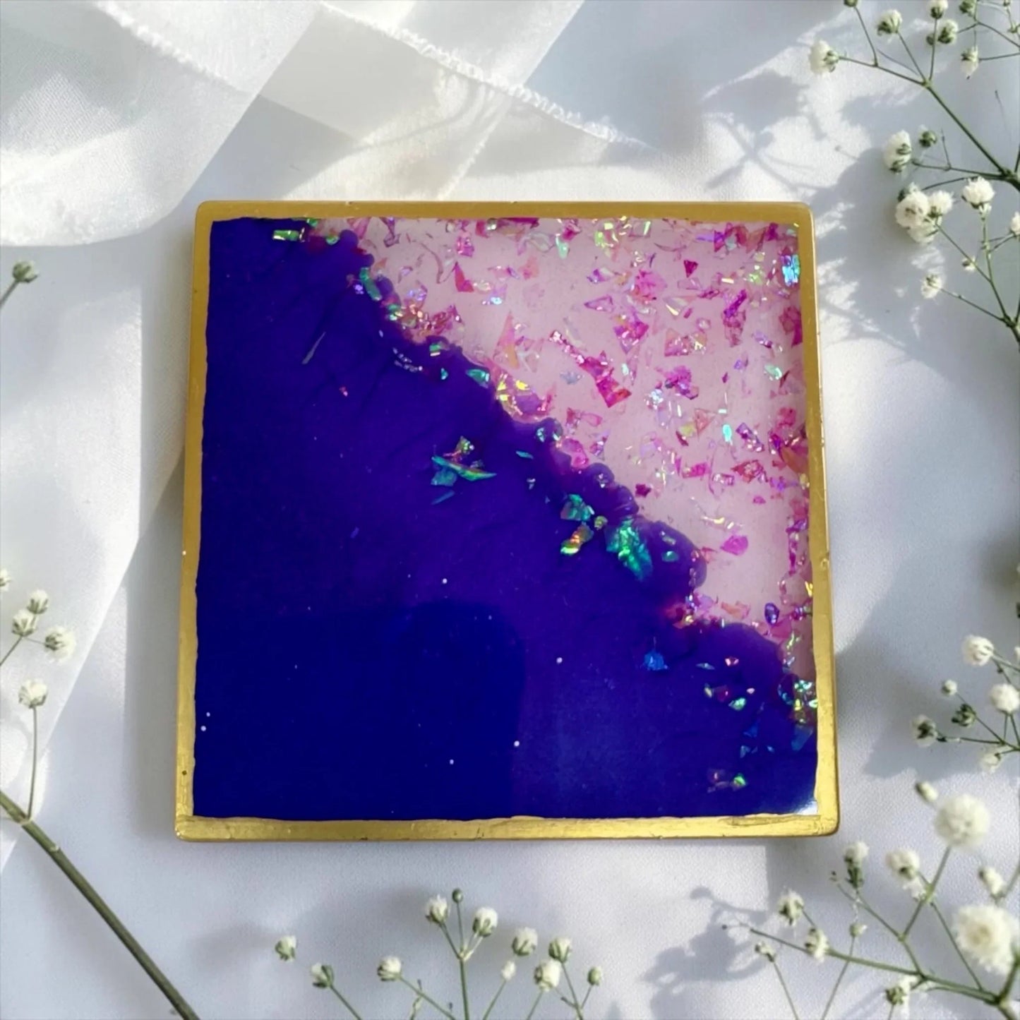 Ein quadratischer Untersetzer aus Epoxidharz in Dunkellila und Rosa mit Goldrand und Glitzerflocken, dekoriert mit weißen Schleierkraut-Blumen.