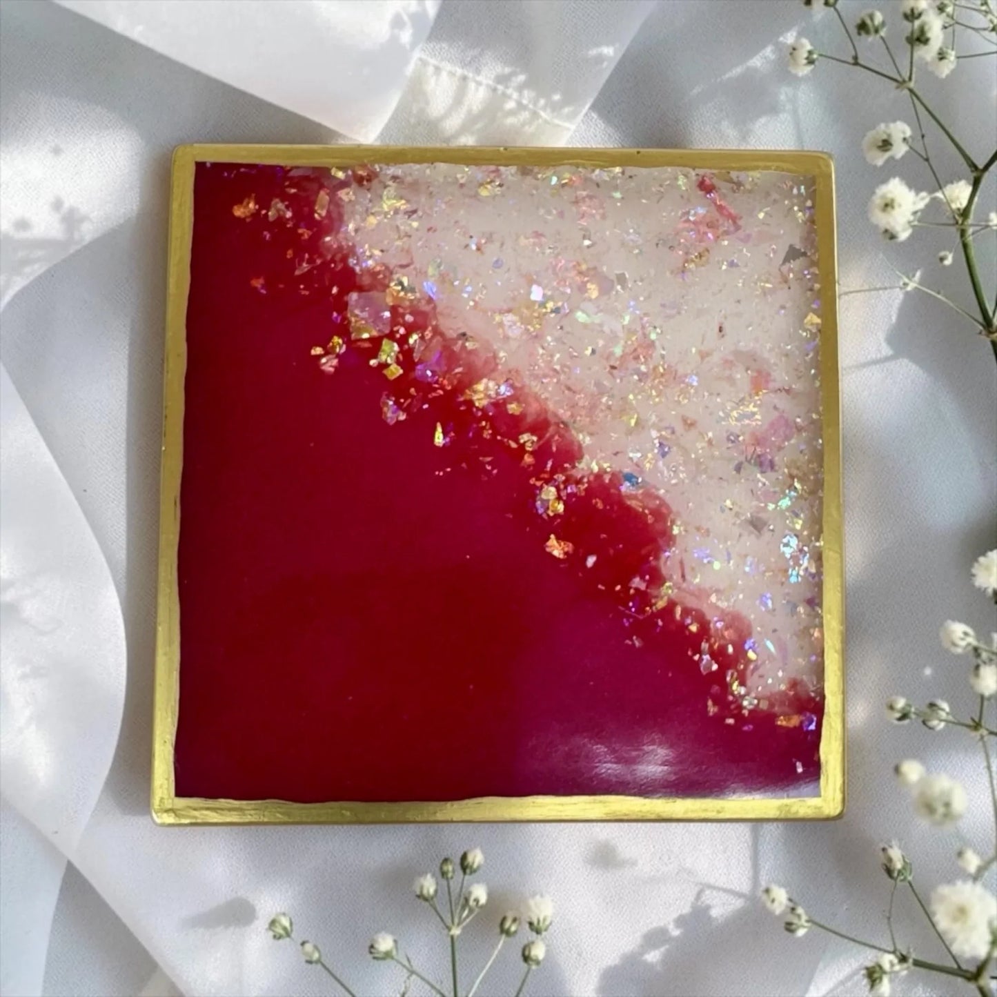 Ein quadratischer Untersetzer aus Epoxidharz in kräftigem Pink und Weiß mit Goldrand und Glitzer, umrahmt von zartem weißen Schleierkraut auf weißem Satin.