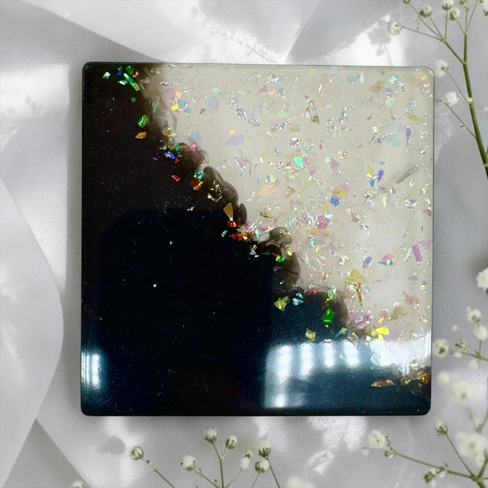 Ein quadratischer Untersetzer aus Epoxidharz in Schwarz und Weiß mit holografischem Glitzer, dekoriert mit weißem Schleierkraut auf hellem Stoff.
