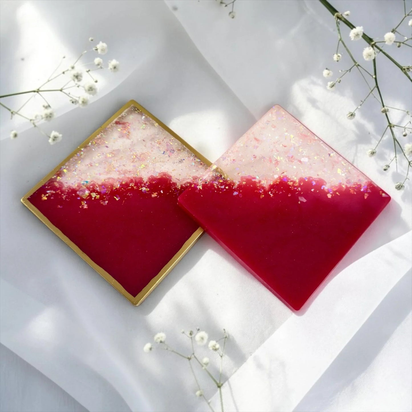 Zwei rautenförmig angeordnete Epoxidharz-Untersetzer in kräftigem Pink und Weiß mit Glitzer, einer mit Goldrand, dekoriert mit weißem Schleierkraut.