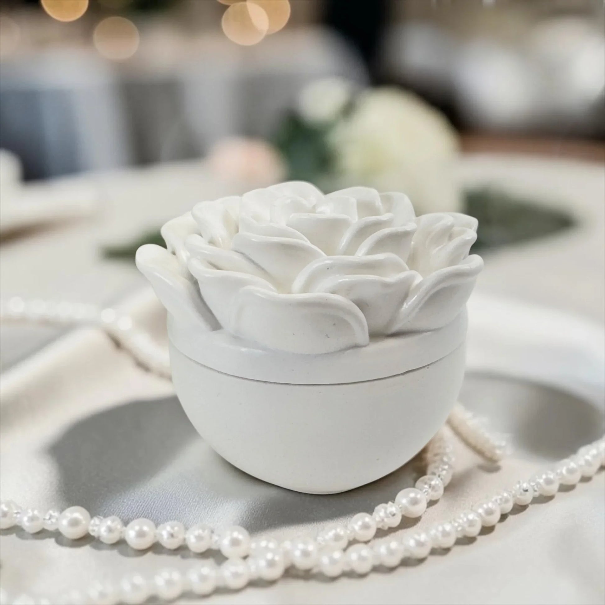 Nahaufnahme einer weißen Keramik-Duftkerze mit Kameliendeckel, elegant dekoriert mit Perlenketten auf einem festlich gedeckten Tisch.